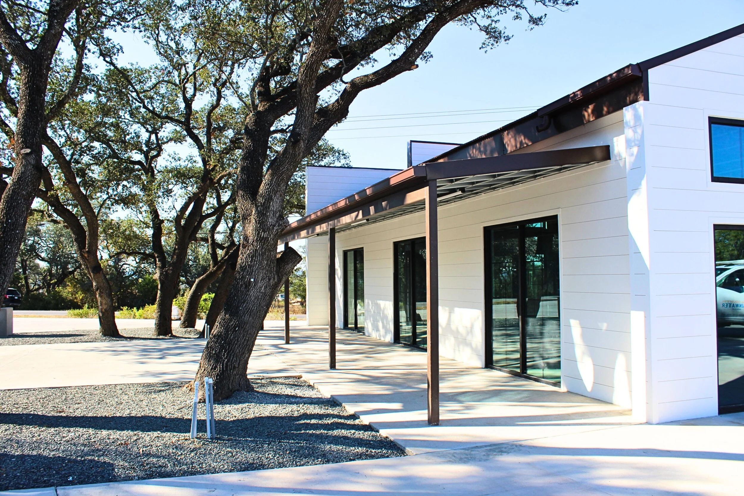 Modern white building with large glass windows and a small porch with black metal posts, surrounded by trees with broad branches, on a sunny day with clear blue sky.