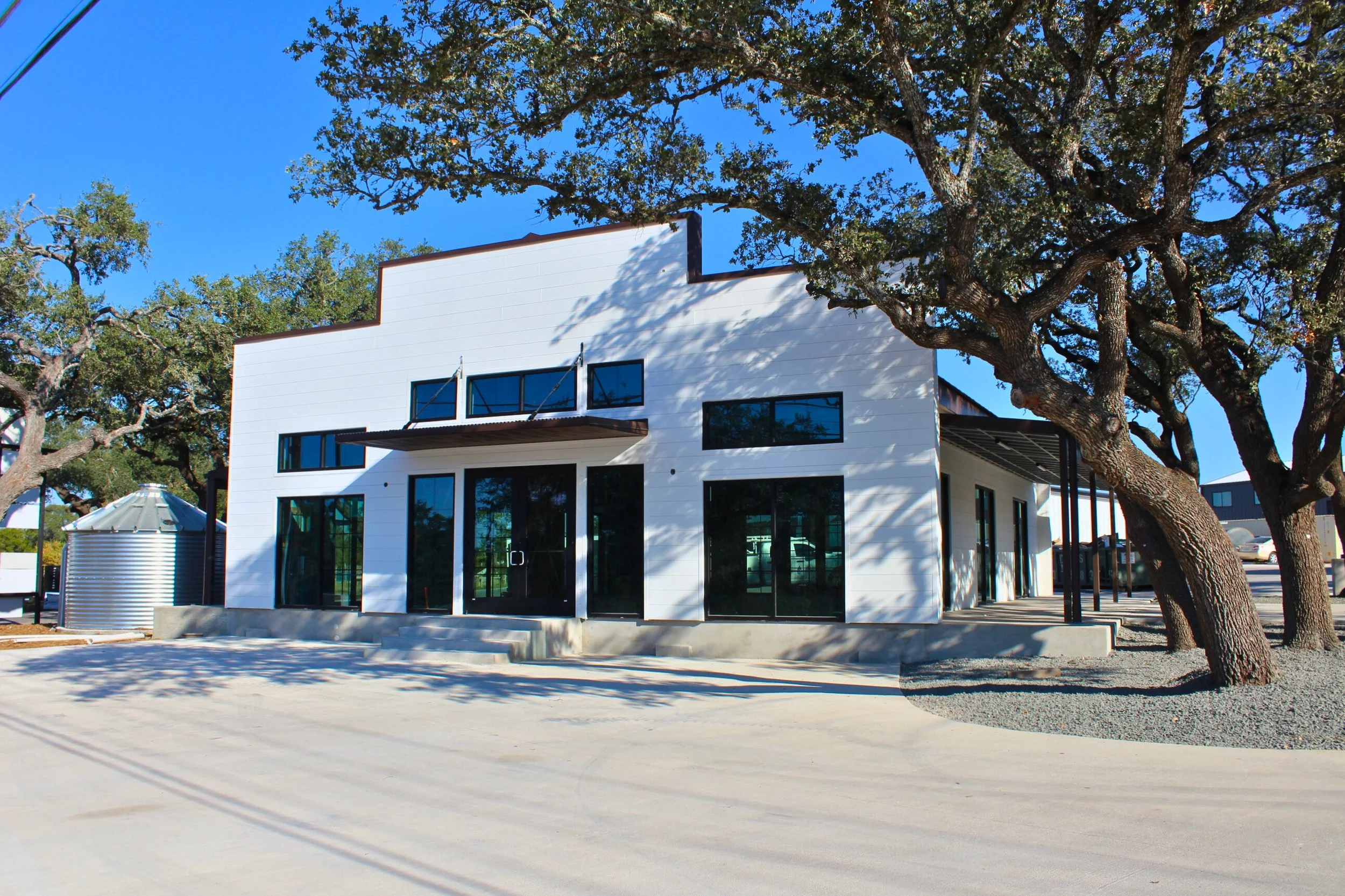 Modern white building with black window frames and sliding glass doors, surrounded by large trees, under a clear blue sky.