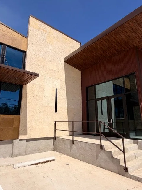 Modern building exterior with beige stone walls, wood accents, large glass windows, and concrete stairs with metal handrails.