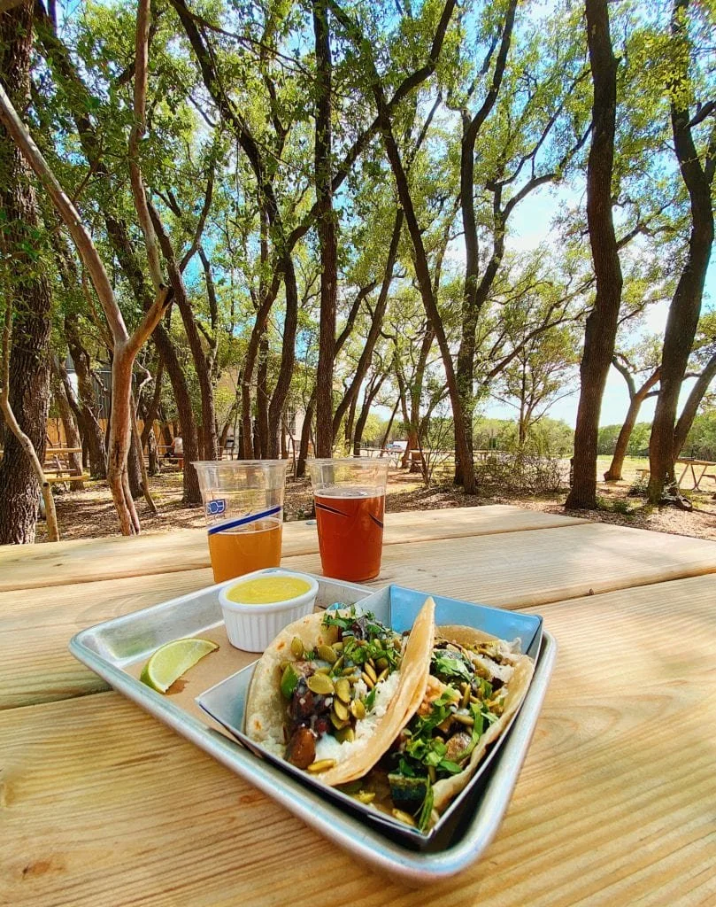 A tray with two tacos, lime wedge, and a small cup of yellow sauce, accompanied by two cups of drinks on a wooden table in a wooded outdoor setting.