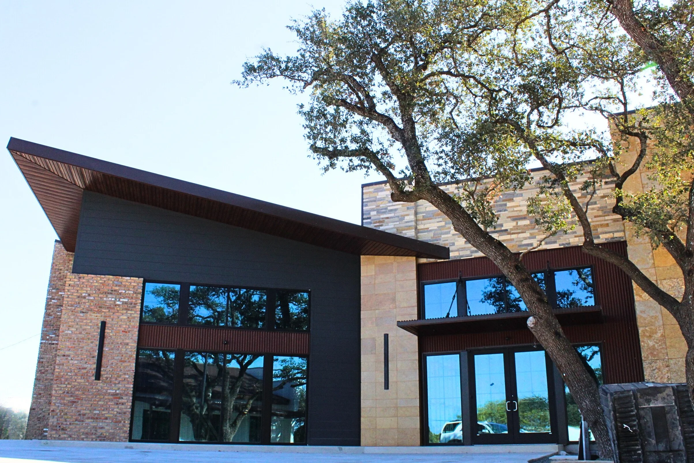 Modern house with brick and dark siding, glass windows, and a large tree in the front yard.