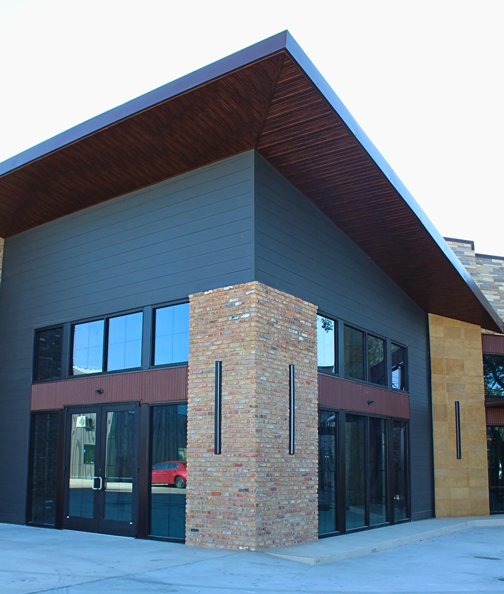 Modern building with brick, dark gray siding, and large glass windows, featuring an overhanging roof with wood interior.