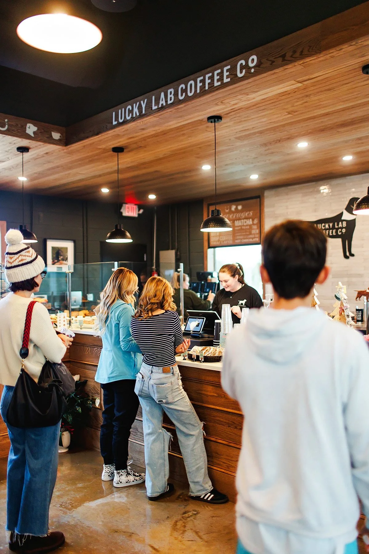 People ordering and picking up coffee at Lucky Lab Coffee Co. in a cozy café with warm wooden accents and hanging pendant lights.