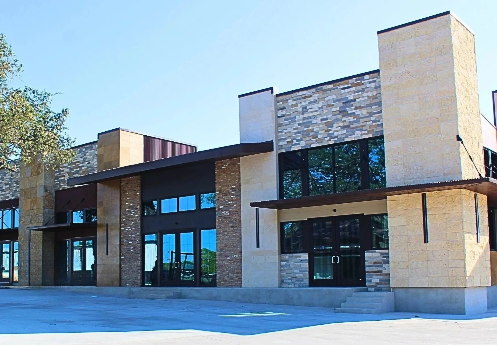 Modern building with a mix of stone, brick, and glass exterior, featuring large windows and a flat roof, set against a clear blue sky.