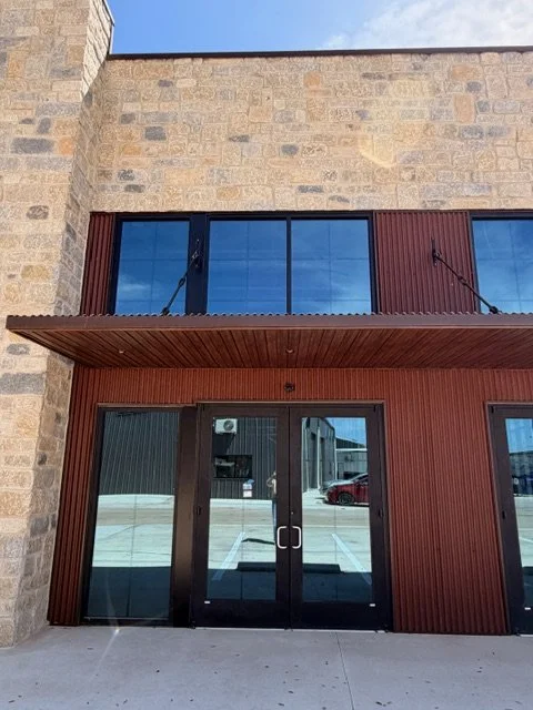 The entrance of a building with glass double doors and brick and wood siding exterior, with reflections of nearby buildings and cars in the glass.