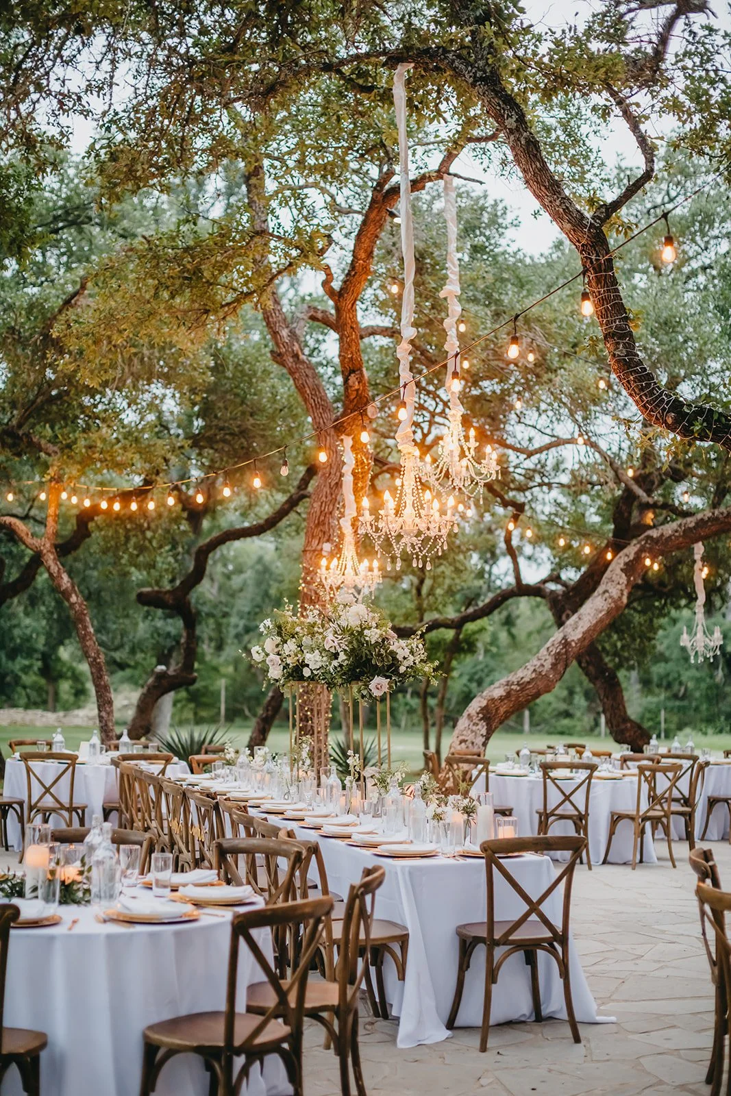 Outdoor wedding reception setup with round tables and wooden chairs, decorated with white tablecloths and floral centerpieces, under string lights and chandeliers hanging from tree branches.