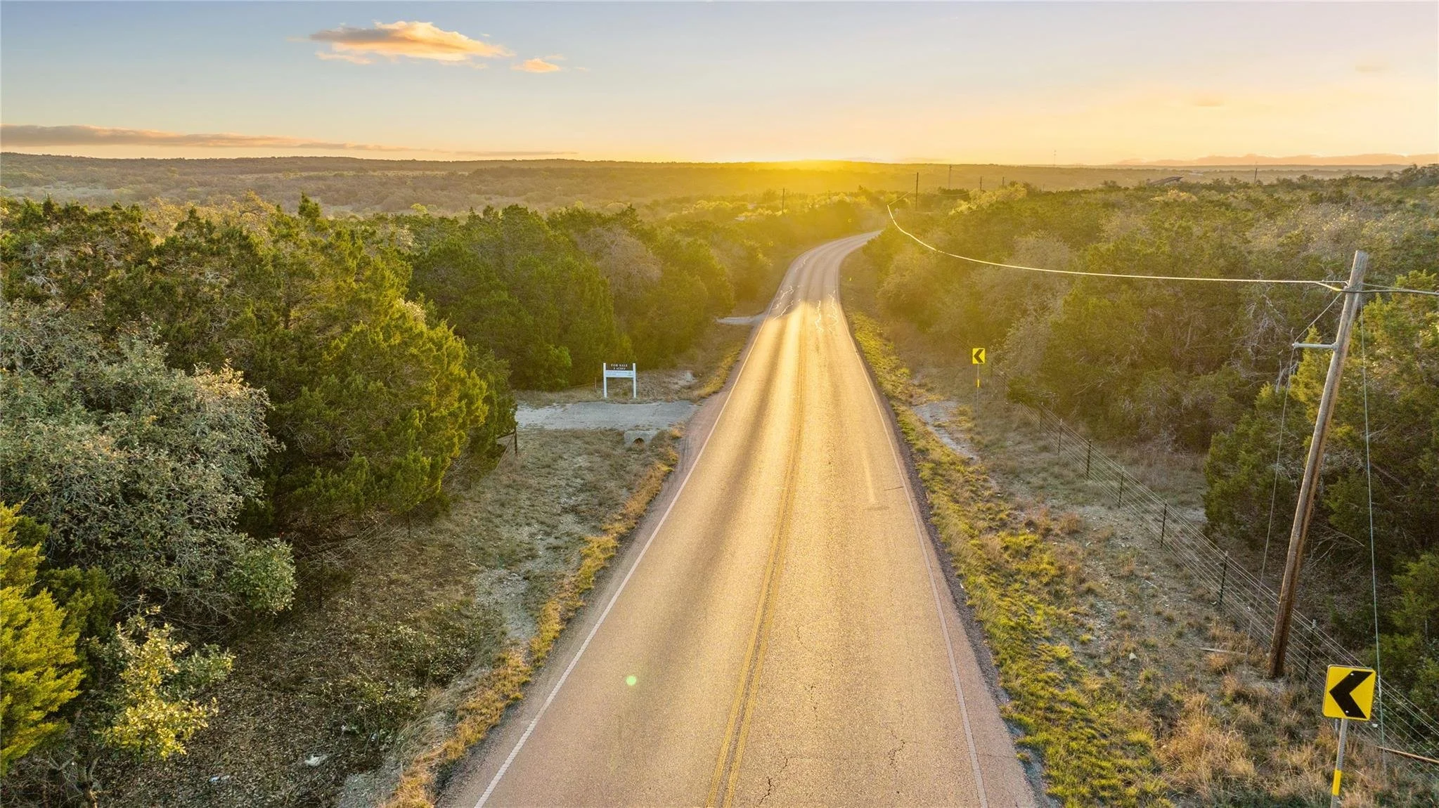 A dirt country road at sunset, surrounded by green trees and shrubbery, with power lines running alongside and a small sign indicating a curve ahead.
