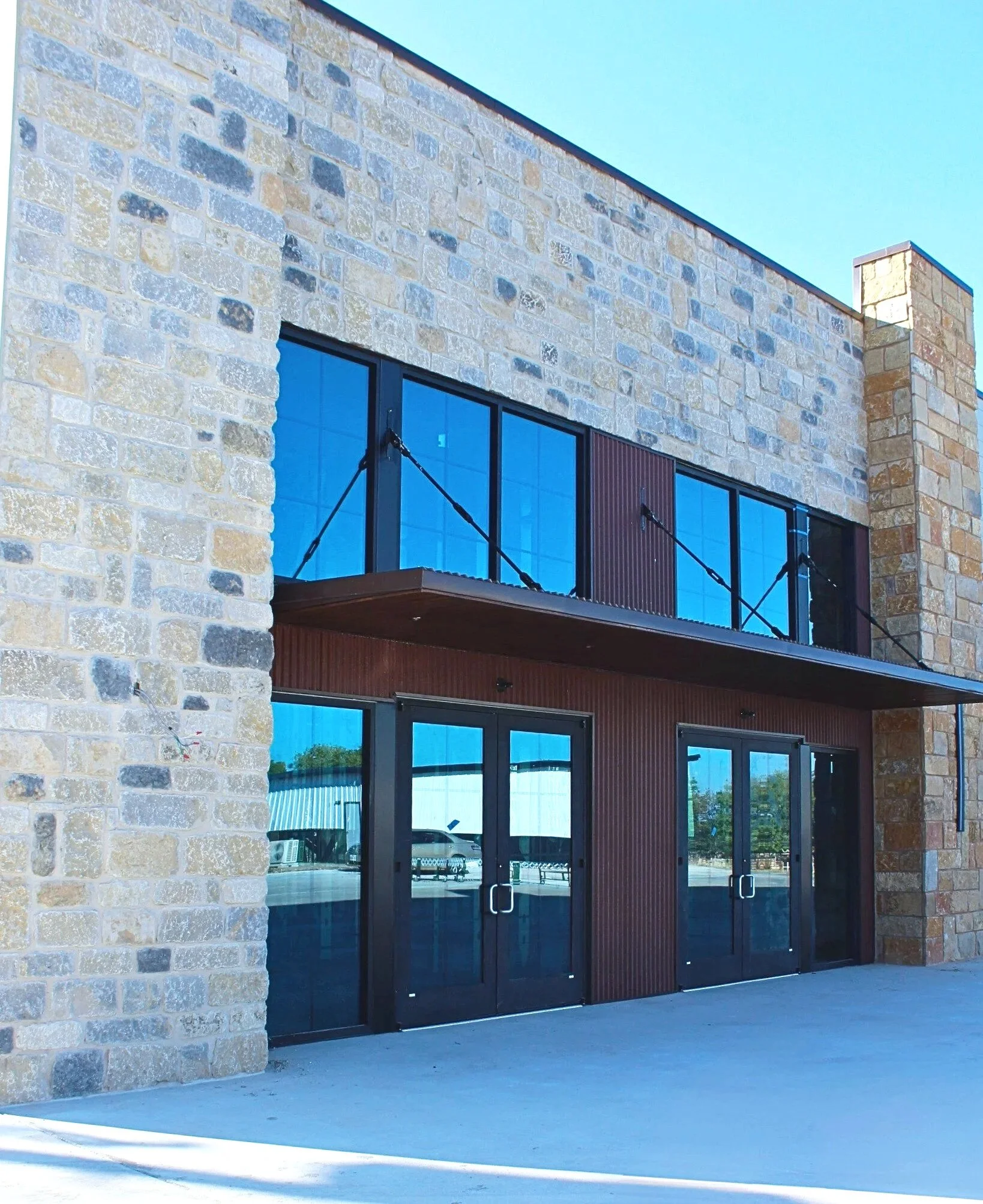 A modern building with large windows and a stone brick facade, featuring a glass door with a metal handle, set against a clear blue sky. The building has a balcony with black metal railing.