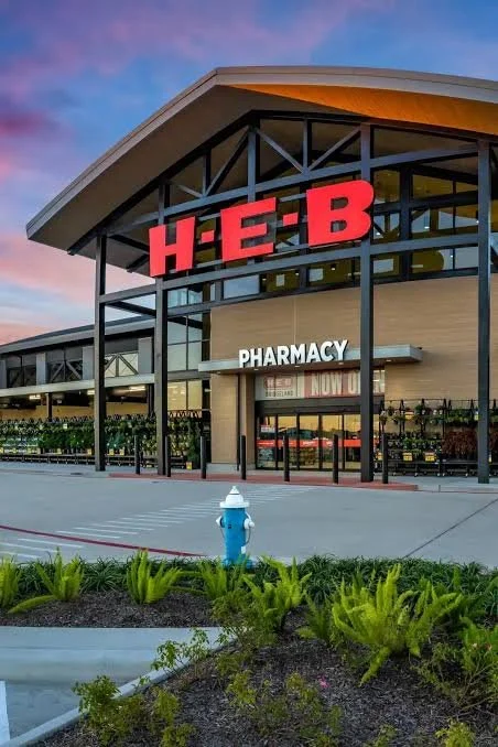 The exterior of an H-E-B grocery store with large red sign, pharmacy entrance, and a landscaped area with plants in the foreground during sunset.