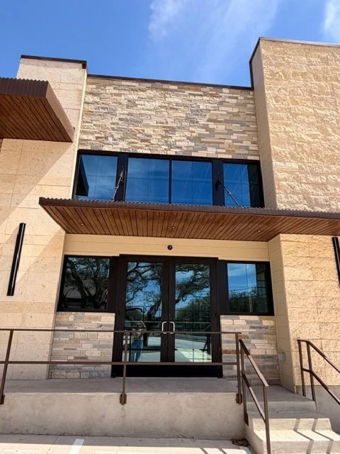 Modern building entrance with glass doors, stone and brick exterior walls, and an overhanging wooden awning, under a blue sky.