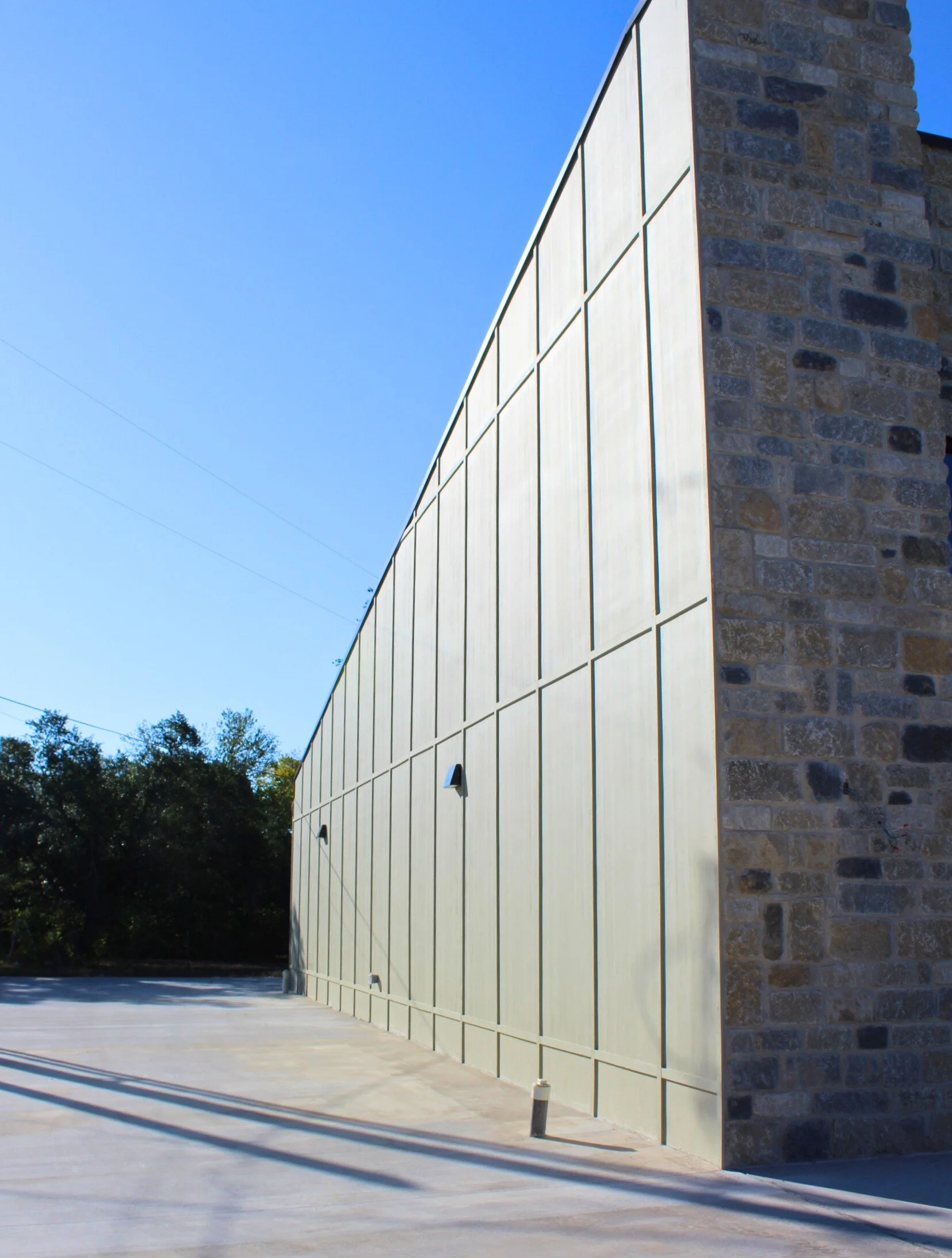 Modern building with a stone wall on the right and a beige metal panel wall on the left, under a clear blue sky, with a concrete sidewalk and parking lot in the foreground.