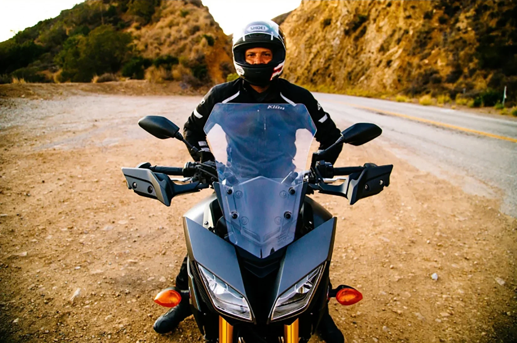 A person wearing a black motorcycle helmet and black riding gear, sitting on a black adventure motorcycle with a transparent windshield, on a dirt trail beside a paved road in a mountainous desert landscape.