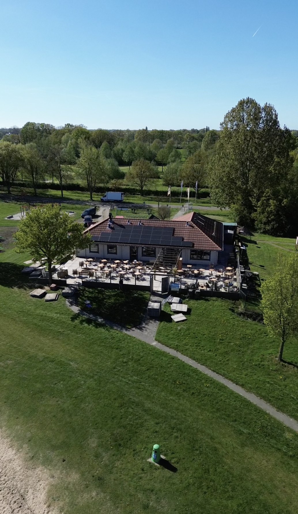 Een gebouw met een terras en muurschermen, omgeven door groene bomen en grasveld, met een helderblauwe lucht erboven.