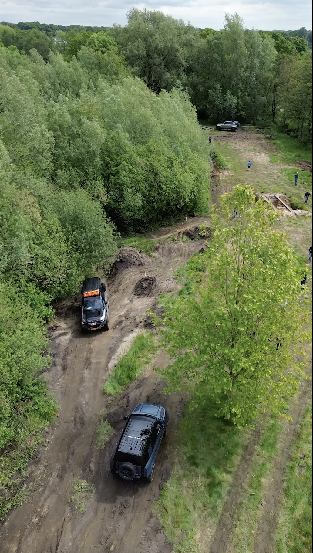 Een bosrijke omgeving met een modderige weg waarop twee zwarte auto's rijden. In de achtergrond zien we een groep mensen en een voertuig geparkeerd onder de bomen, met een grijze hemel erboven.