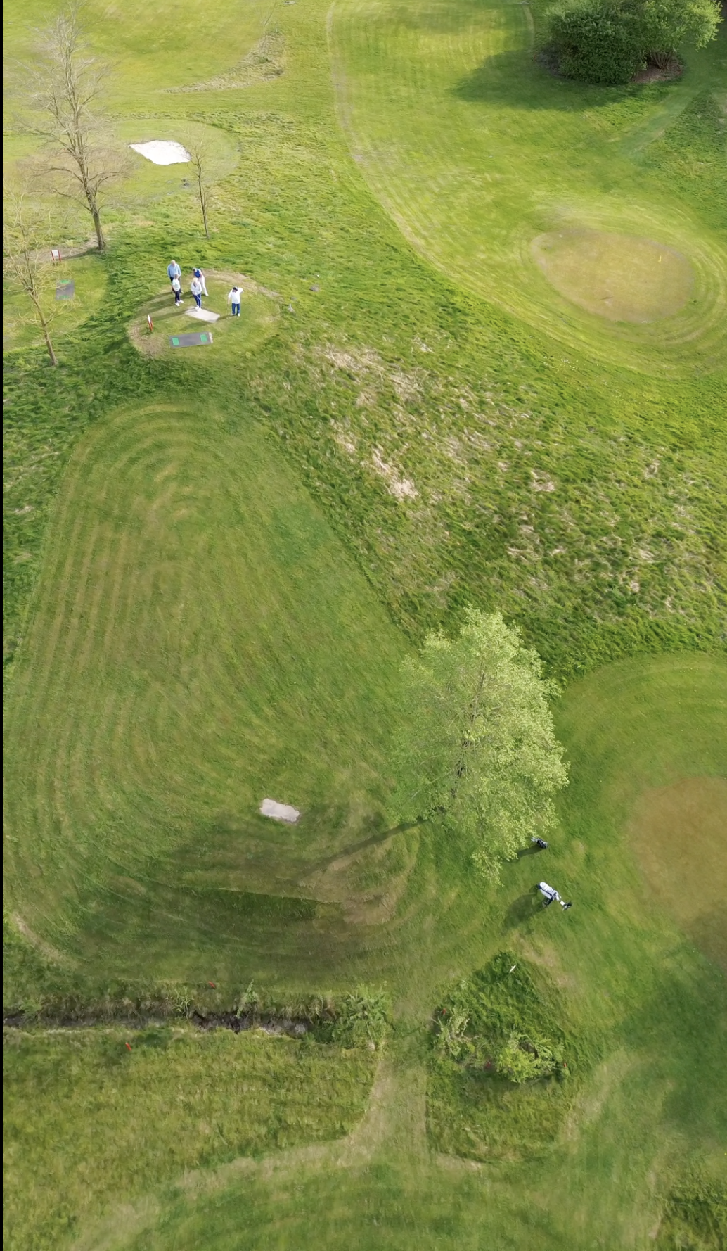Luchtfoto van een golfbaan met enkele golfspelers die aan het oefenen zijn, omringd door groene bomen en grasvelden.