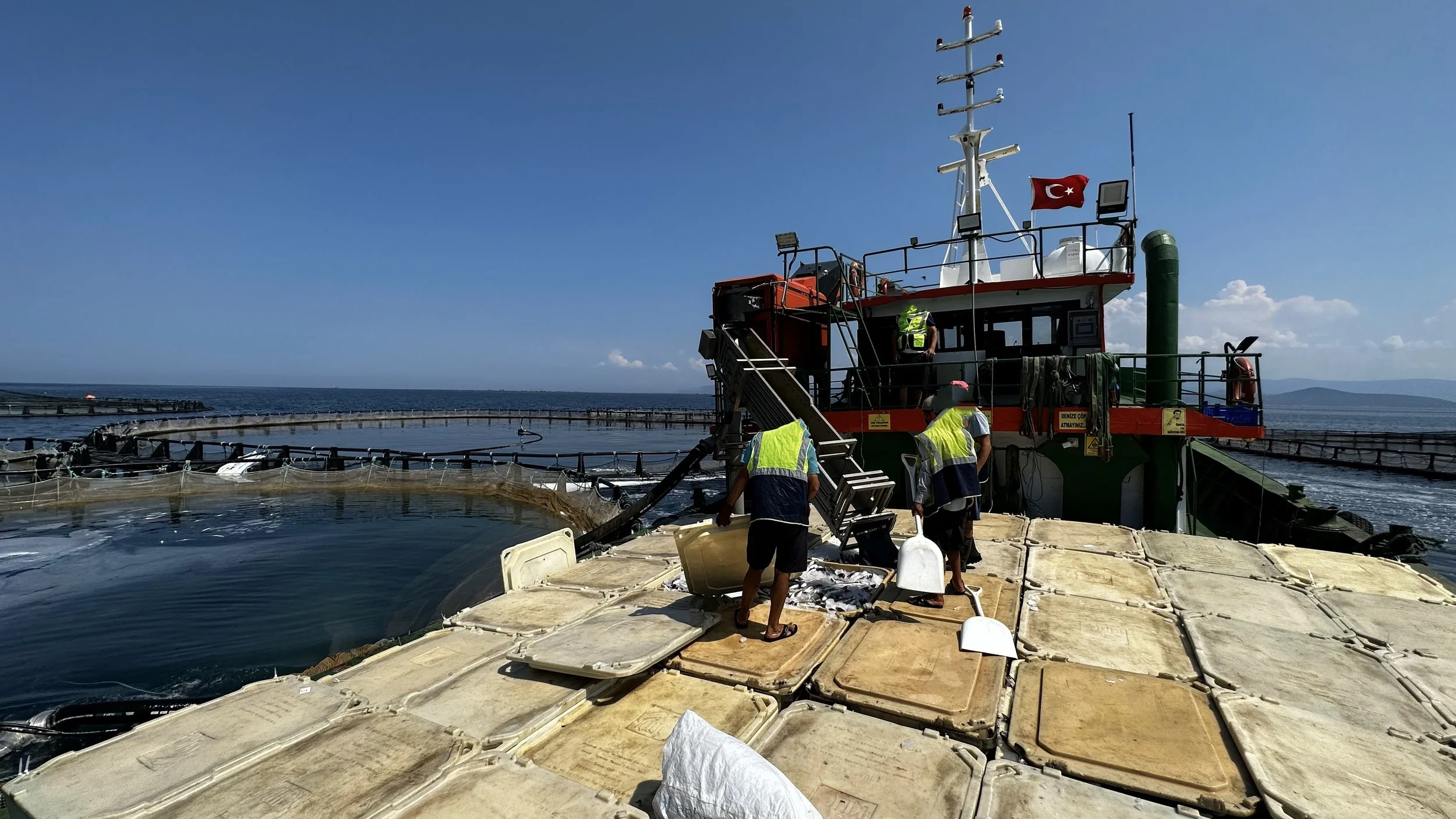 Denizde çalışan insanlar, bir gemi veya platform üzerinde çalışma yapıyor. Görevli kişiler yüksek görünümde, çalışma kıyafetleri ve güvenlik ekipmanlarıyla, ve deniz manzarası arka planda.