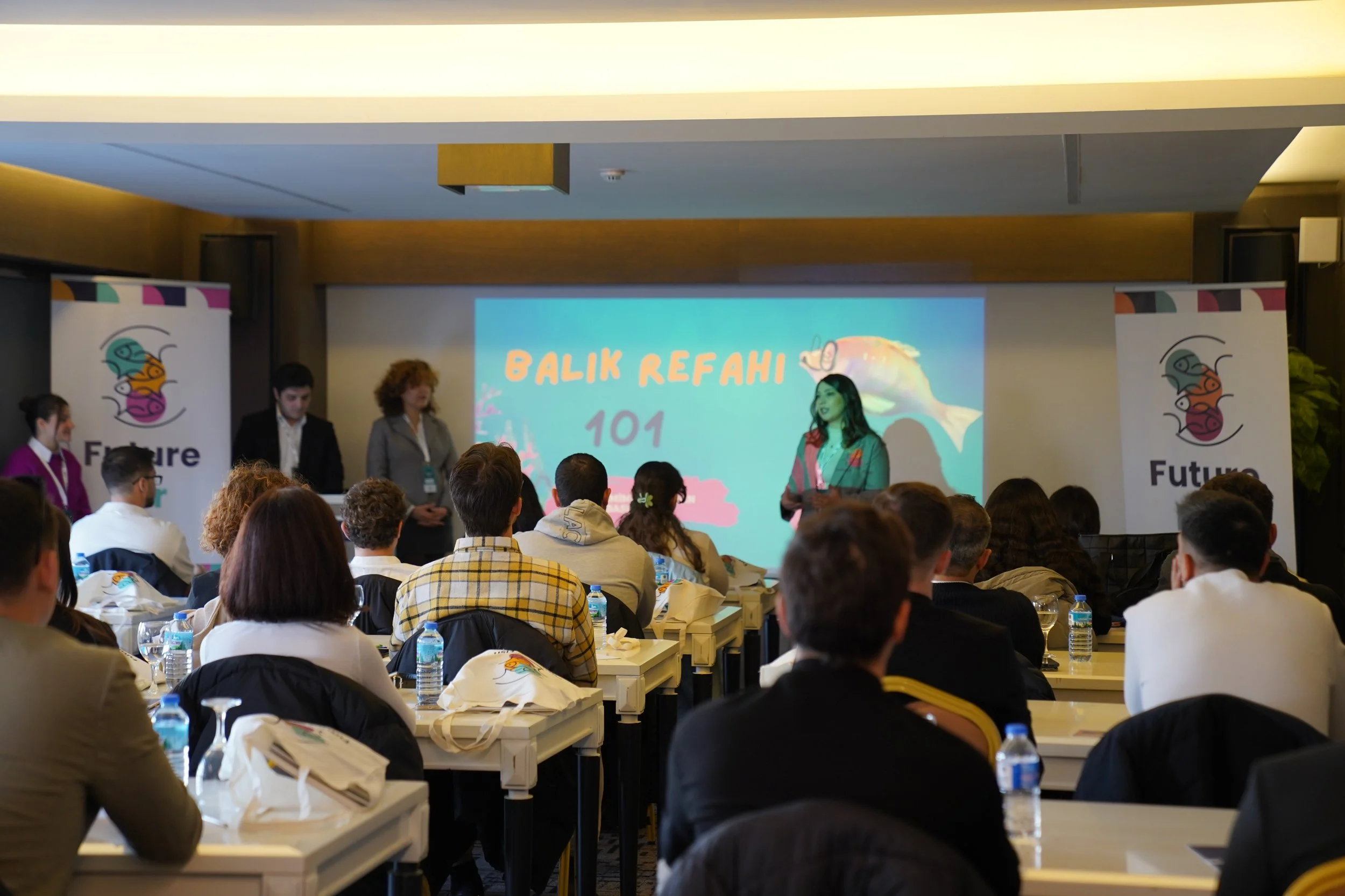 A conference or seminar with attendees sitting at tables, facing a stage with a large screen displaying 'BALIK REFAHI 101'. There are banners on either side with a colorful fish logo and the word 'Future' beneath the logo.