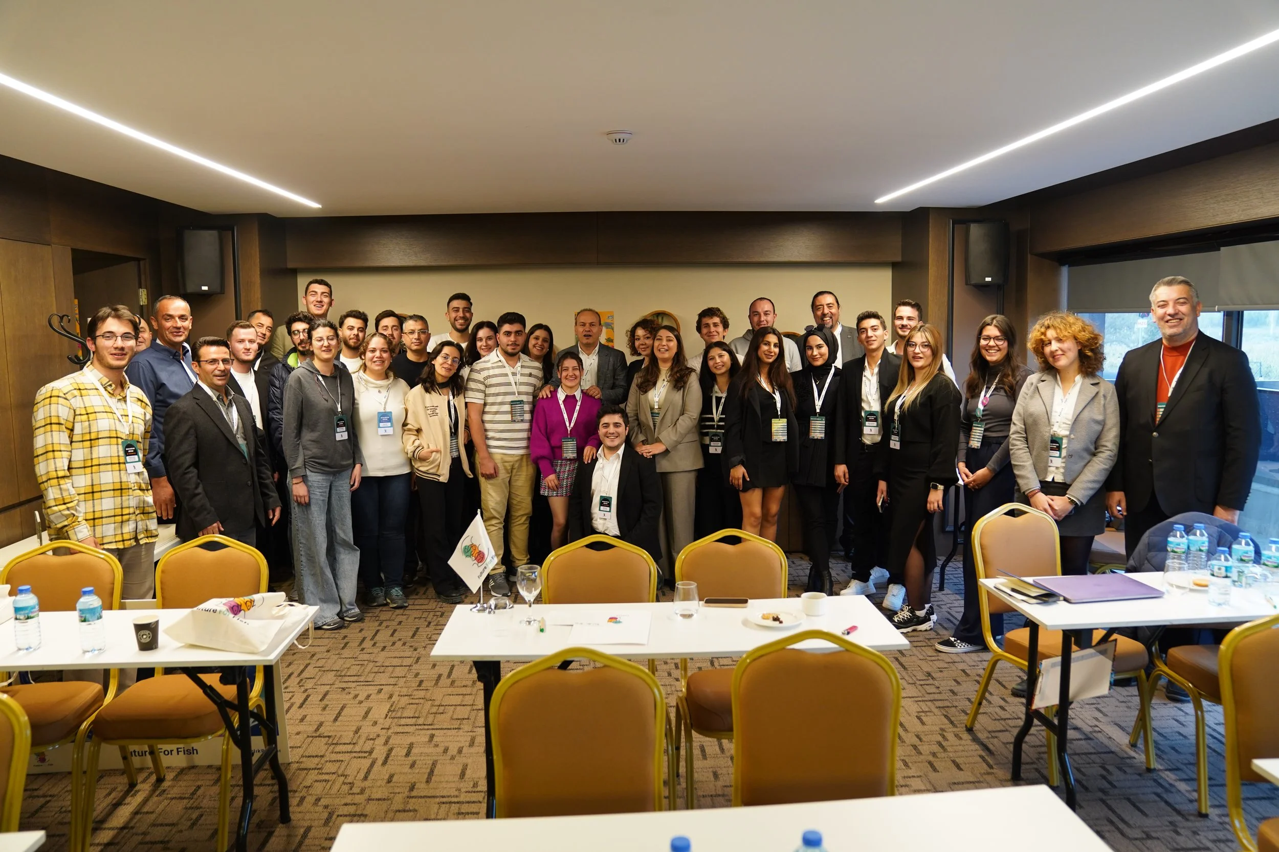 Toplamda 26 kişi, bir toplantı odasında grup fotoğrafı çekiyor. Masada su şişeleri ve not defterleri var. Oda duvarlarında küçük tablolar ve büyük pencereler bulunuyor.