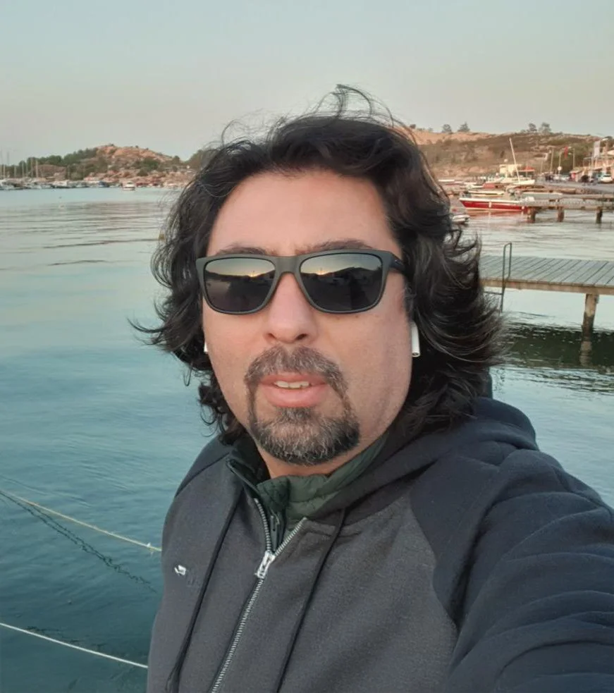 Deniz kenarında, turuncu ve beyaz teknelerin olduğu bir iskelede, erkek bir kişi selfie çekiyor, arka planda deniz ve tepeler görülüyor.