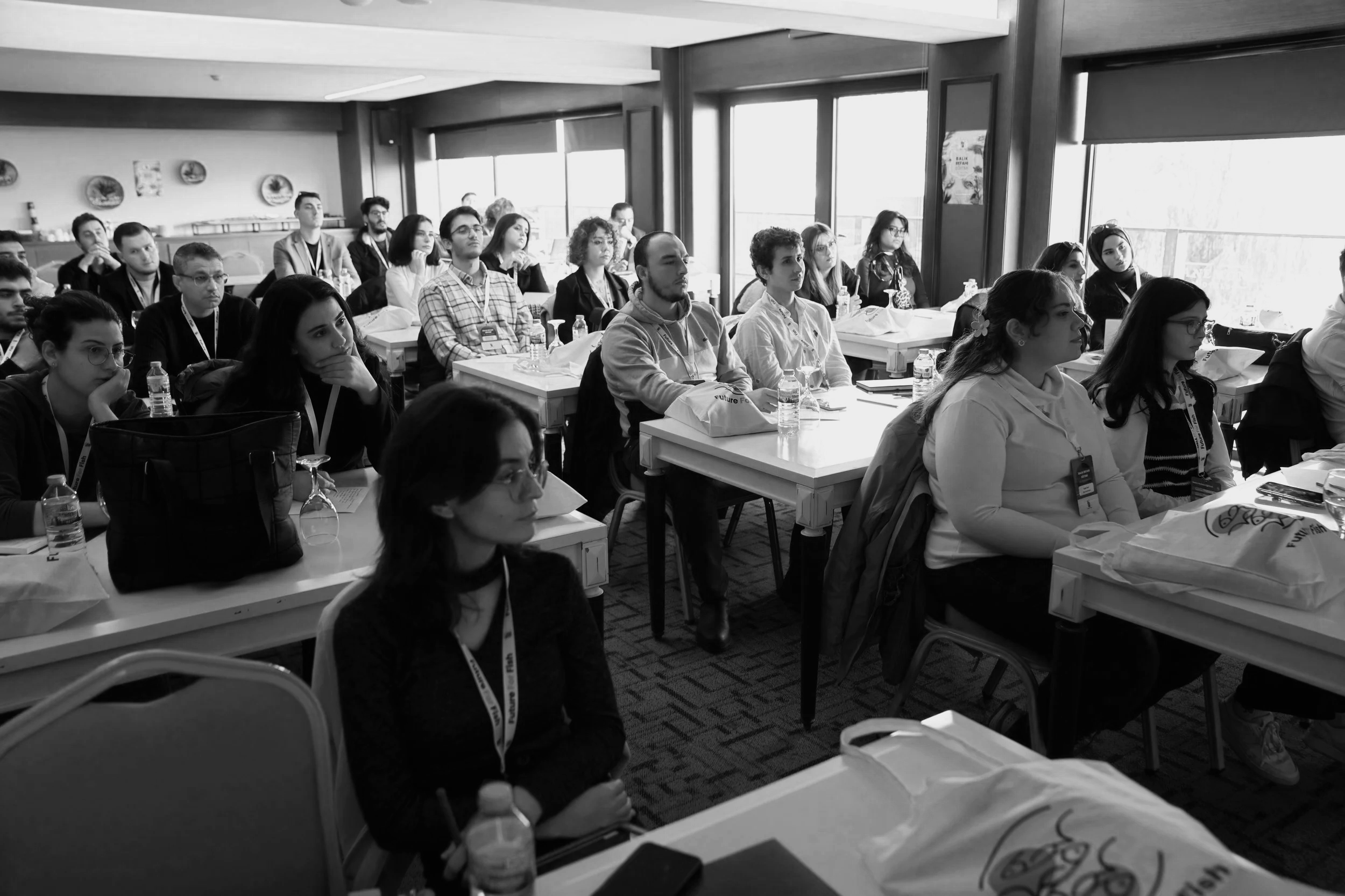 A group of people attending a conference or seminar in a bright room with large windows. They are seated at tables, paying attention to a speaker or presentation. Bottles of water and some conference materials are on the tables.