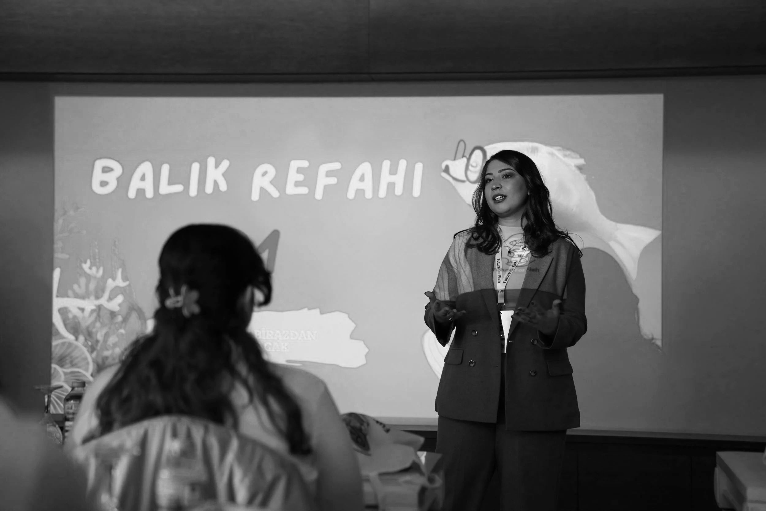 A woman speaking in front of a projection screen with the text 'Balik Rehavi' and a drawing of a whale. The woman has dark hair, wears a blazer, and gestures with her hands. Audience members can be seen in the foreground. The scene appears to be a presentation or lecture.