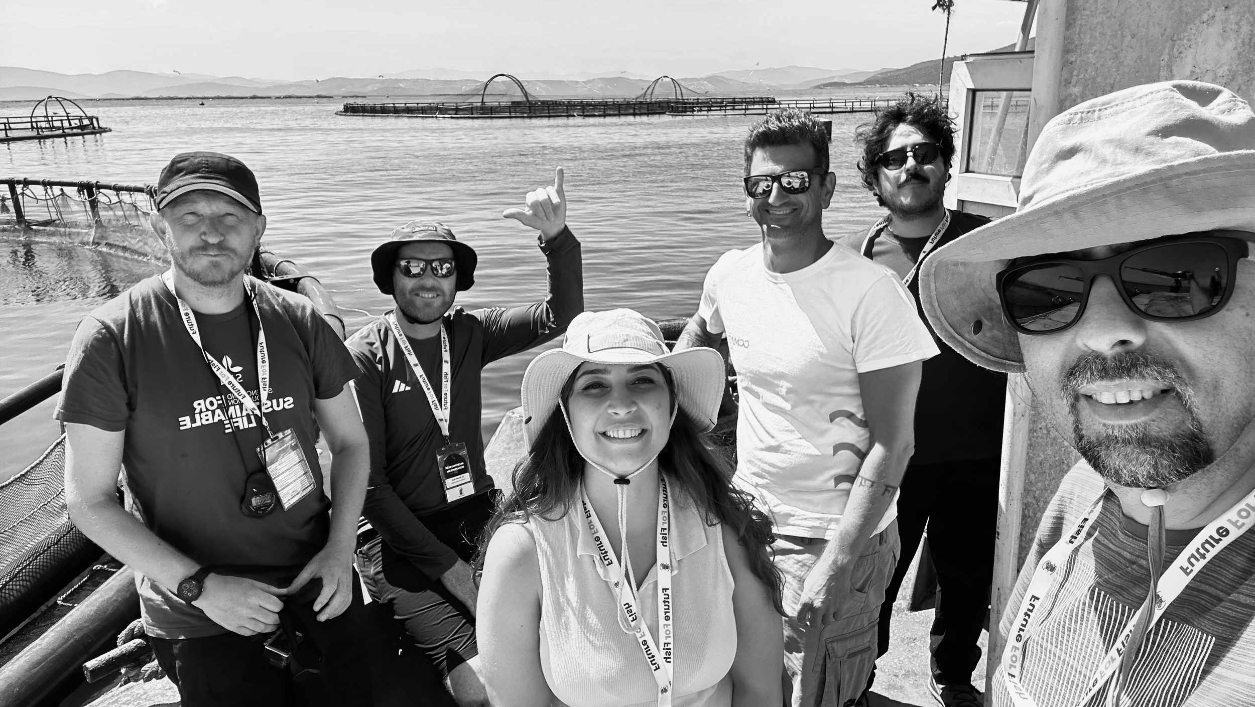 Görüntüde yedi kişi, giyisilerinde kimlik kartları ve şapka veya güneş gözlüğü takmış, göl kenarında bir teknede selfie yapıyorlar. Arka planda su ve uzaklarda dağlar ile iskeleler gözüküyor.