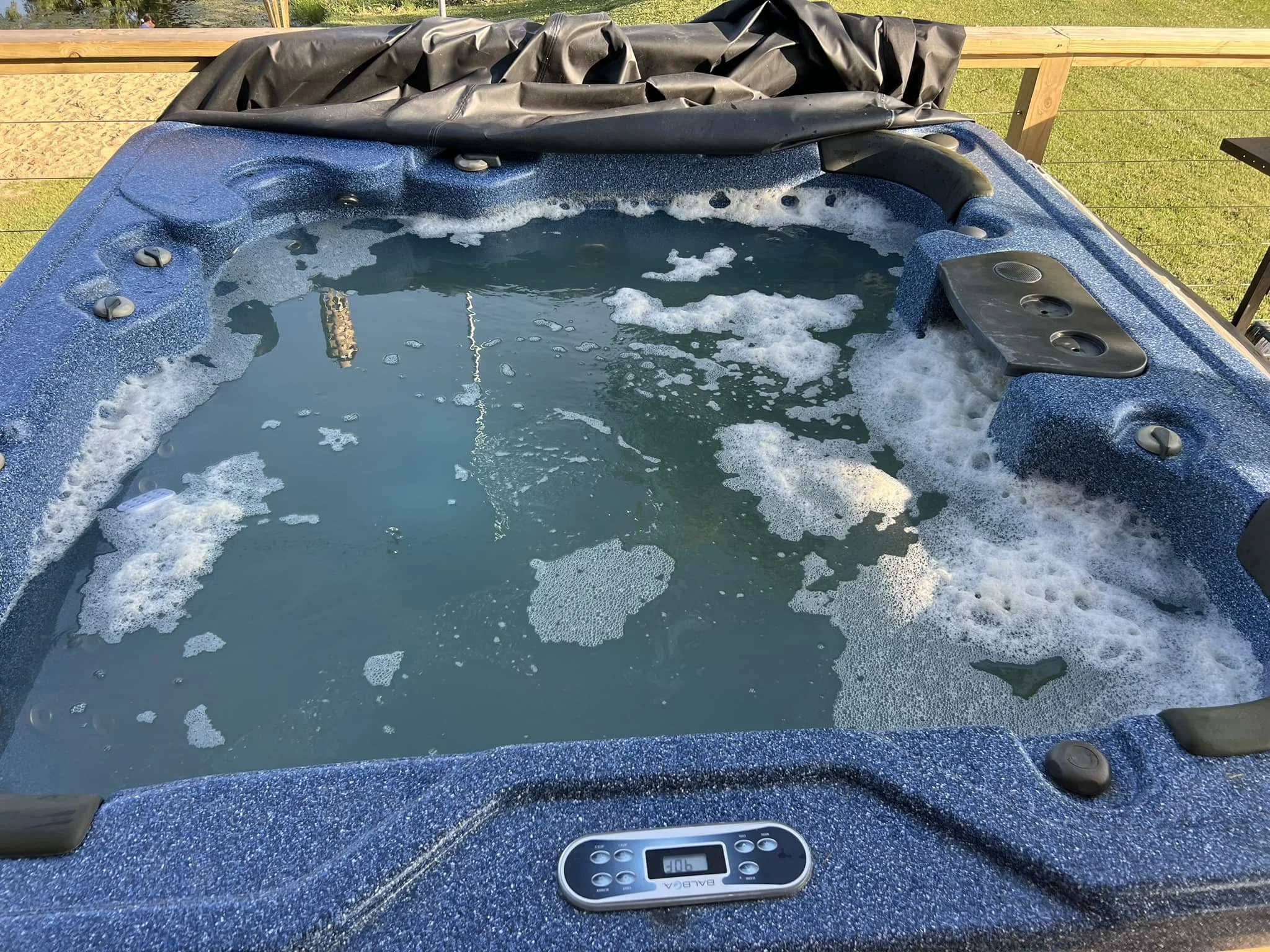 A hot tub with blue speckled exterior filled with water and foam, with black control panel, located outdoors on a wooden deck with grass and a fence in the background.