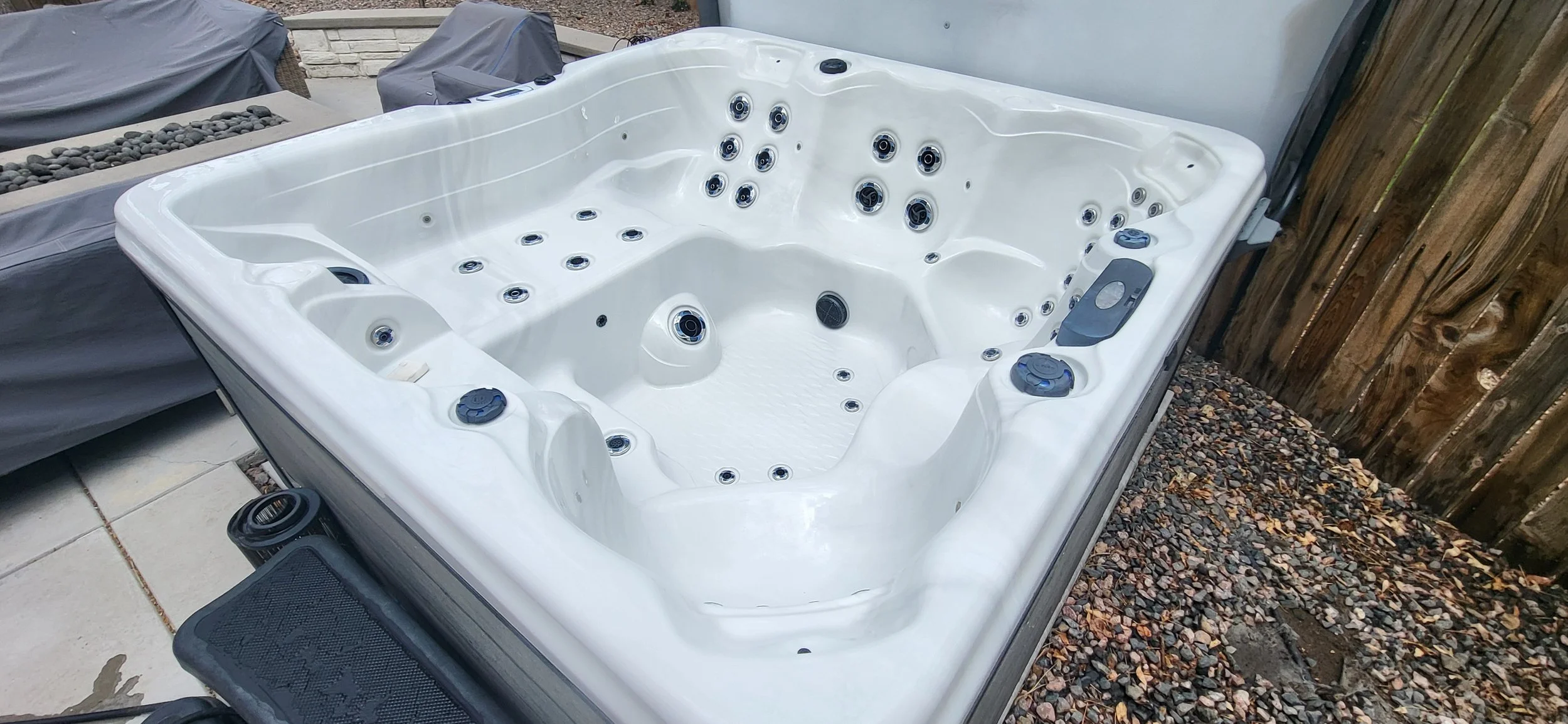 Empty white hot tub with multiple jets, controls, and features outdoors on a patio, next to a wooden fence.
