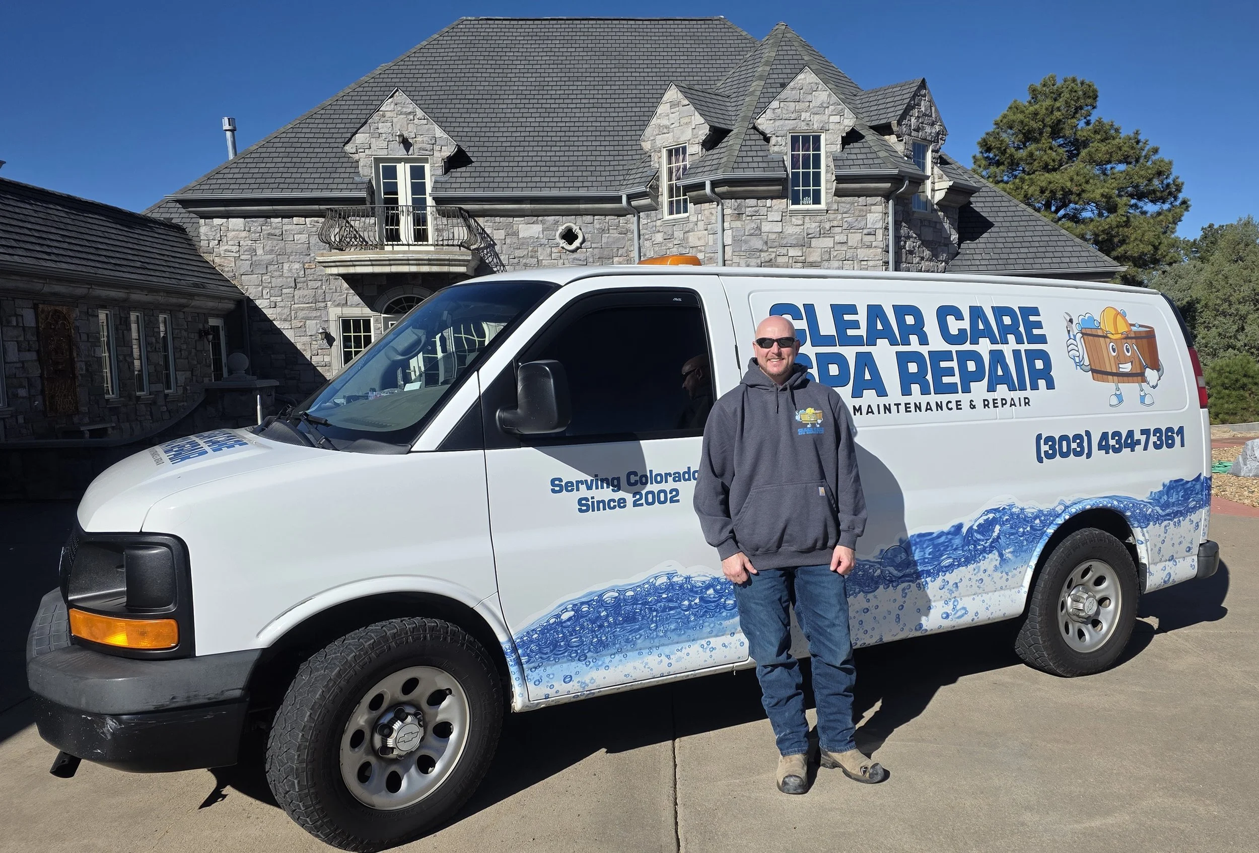 A man stands in front of a white service van that has 'Clear Care Spa Repair' written on it, parked in front of a large stone house with multiple windows and a balcony. The scene is outdoors on a clear, sunny day.