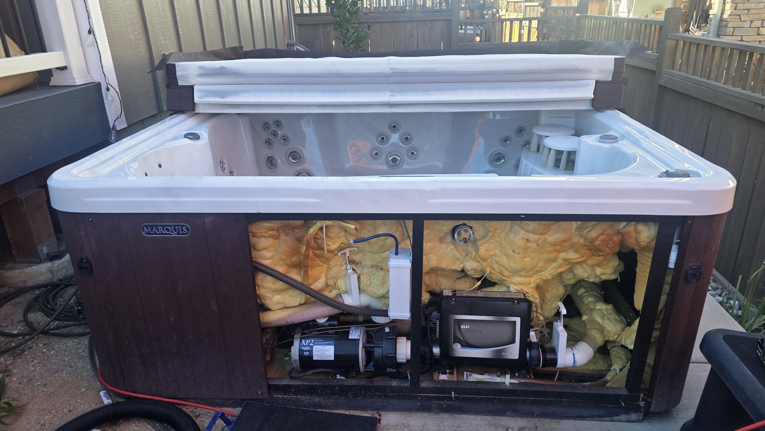 Outdoor hot tub with jets and a partially open cover on a backyard deck, with visible insulation and mechanical components underneath.