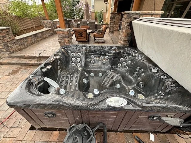 Black and gray hot tub on a brick patio in backyard with outdoor seating area and stone walls in the background.