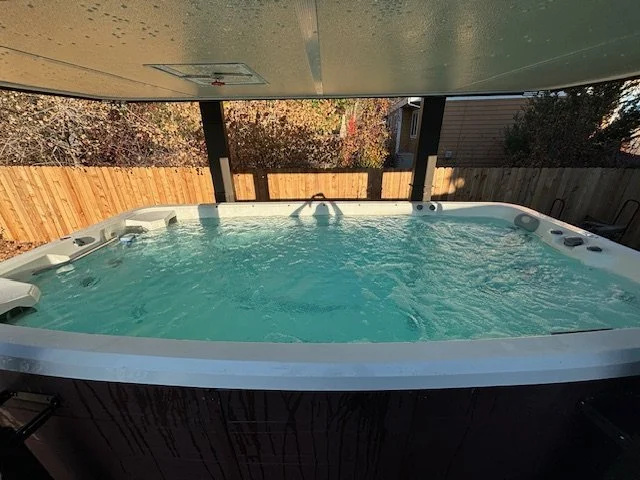 An outdoor hot tub with blue water, surrounded by a wooden fence with trees and a house in the background, under a covered patio.