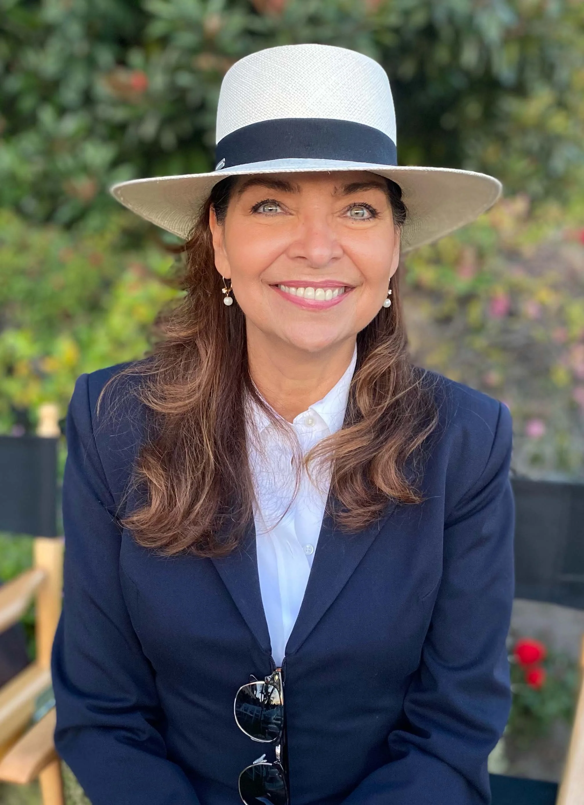 Paige Junaues smiling outdoors, wearing a large white hat with a black ribbon, a navy blazer, white shirt, and pearl earrings, with greenery in the background.