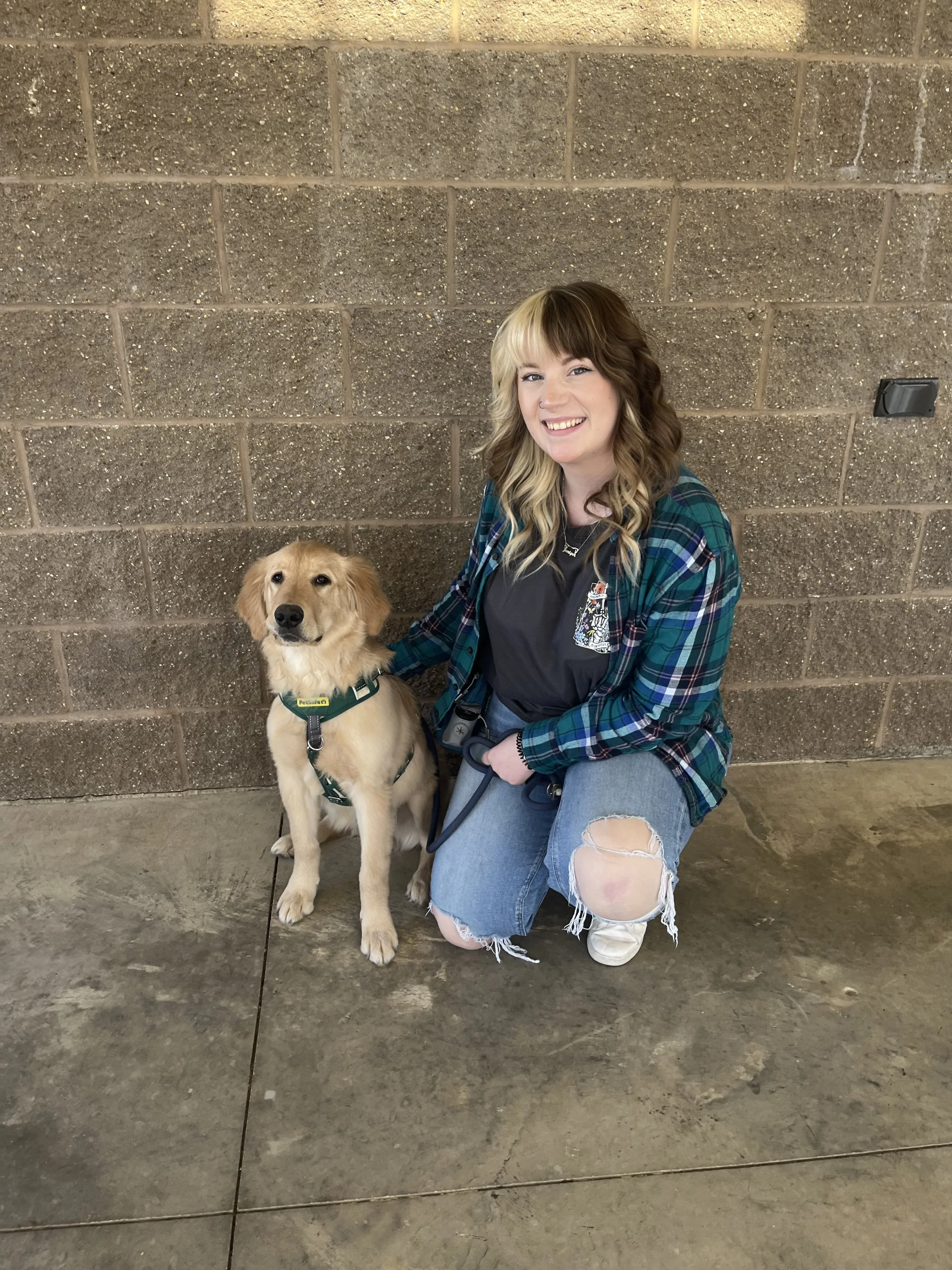A young woman with wavy, half-blonde and half-brown hair kneeling next to a golden retriever puppy against a brick wall.