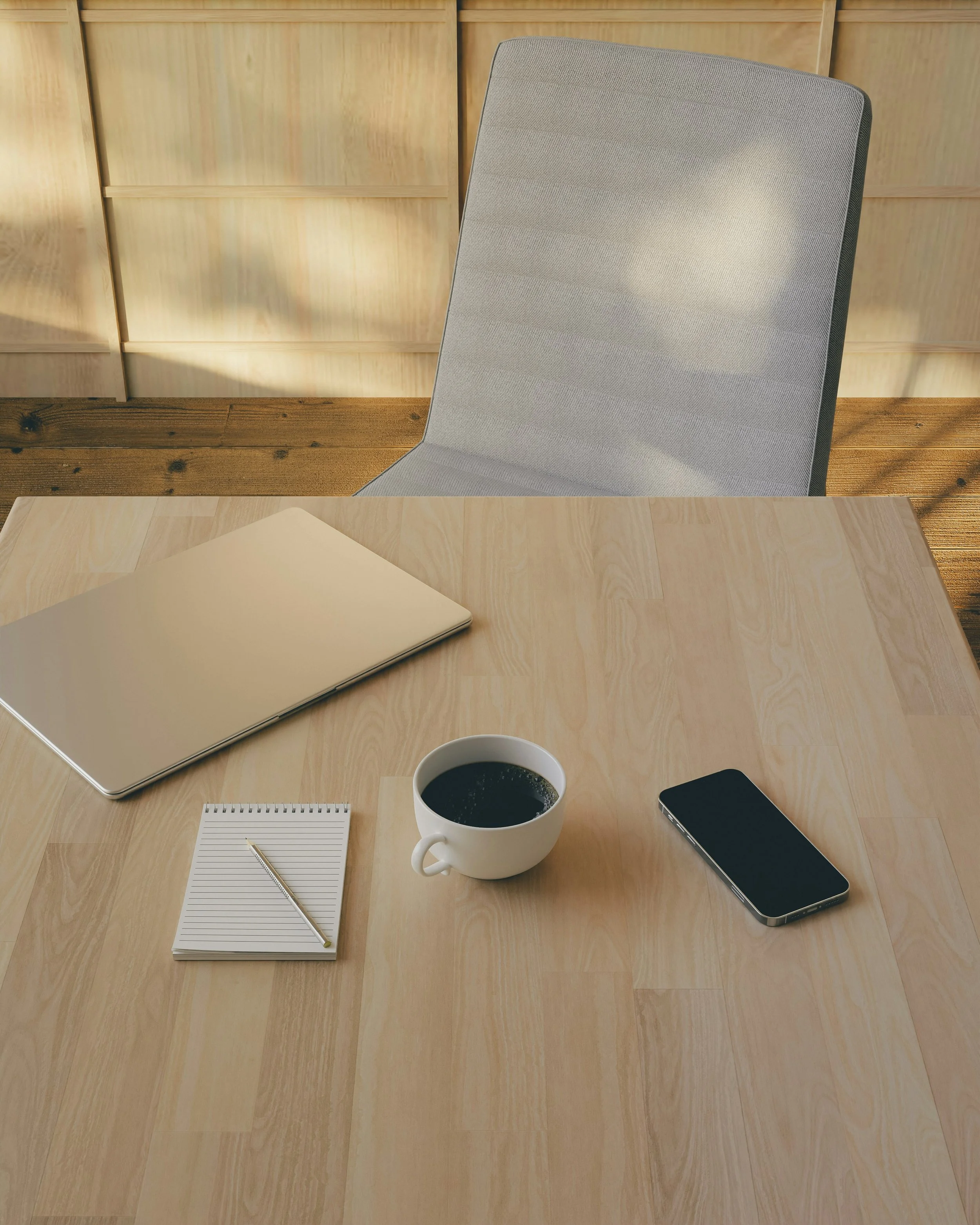 Ein Schreibtisch mit einem Laptop, einer Notizblock, einem Stift, einer Tasse Kaffee und einem Smartphone. Im Hintergrund ein Stuhl und eine Holz-Wand.