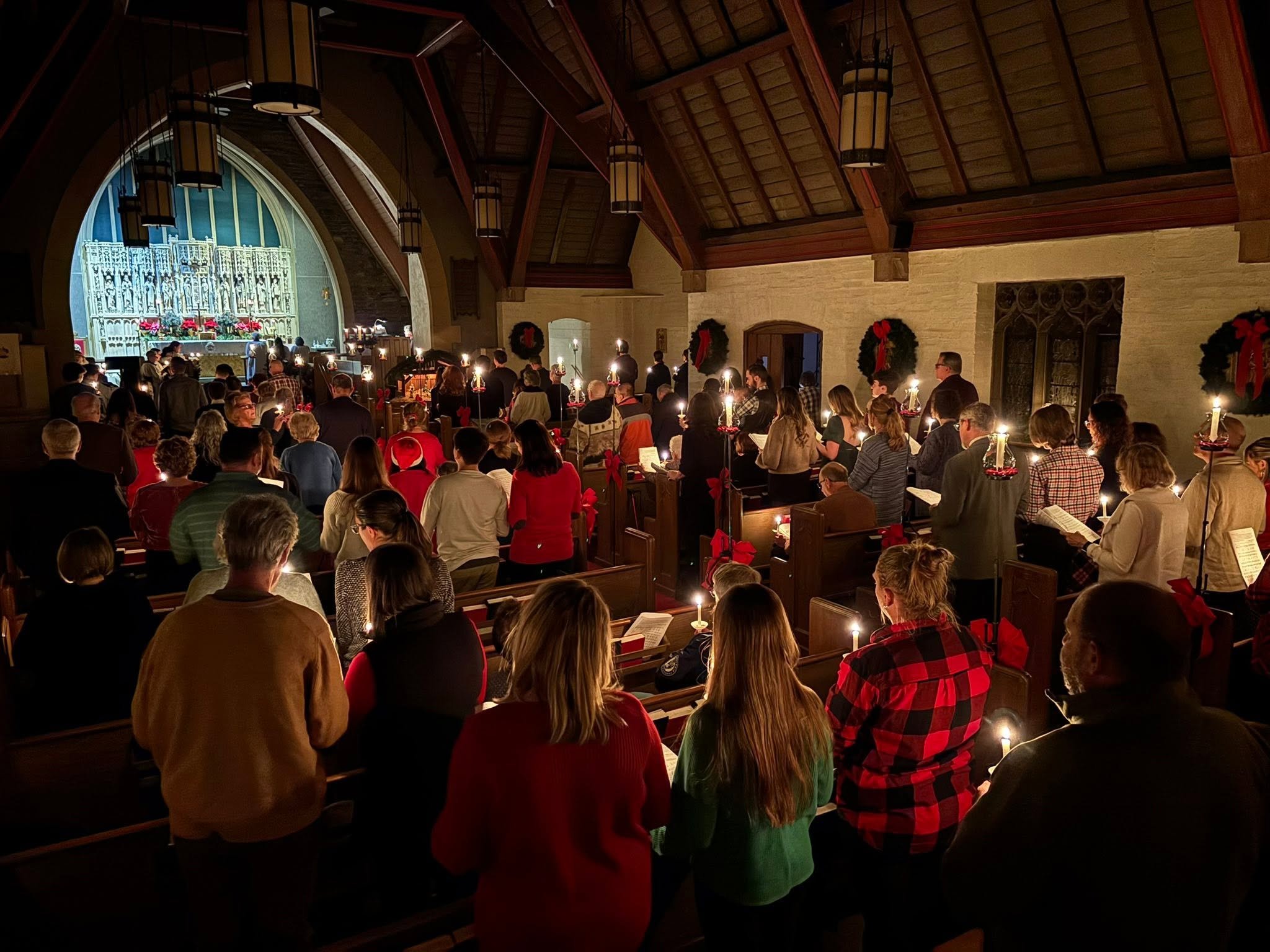 A congregation of people inside a church during a Christmas service, holding candles, with wreaths and festive decorations around.