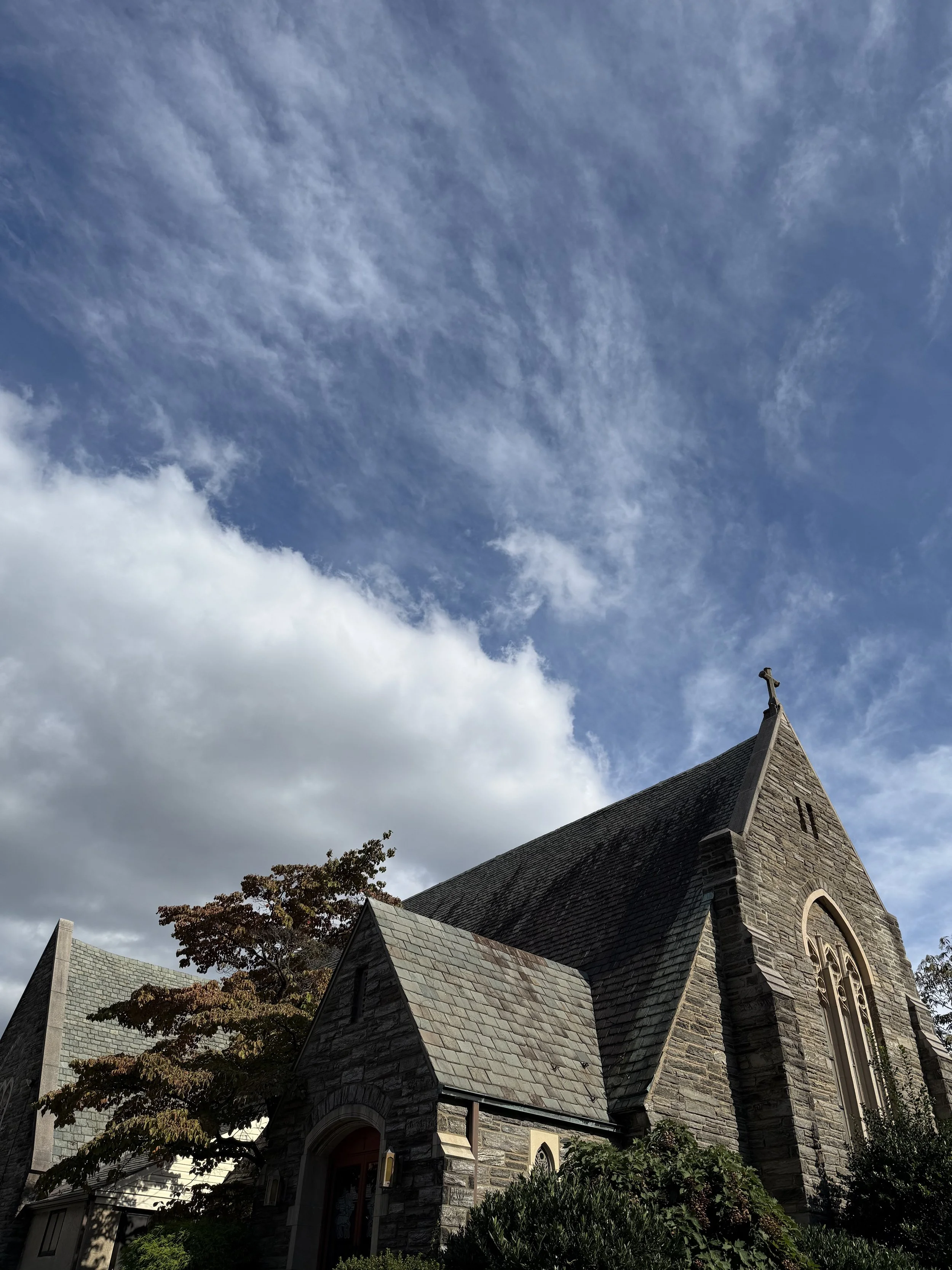 A stone church with a tall steeple and arched windows, surrounded by trees under a partly cloudy sky.