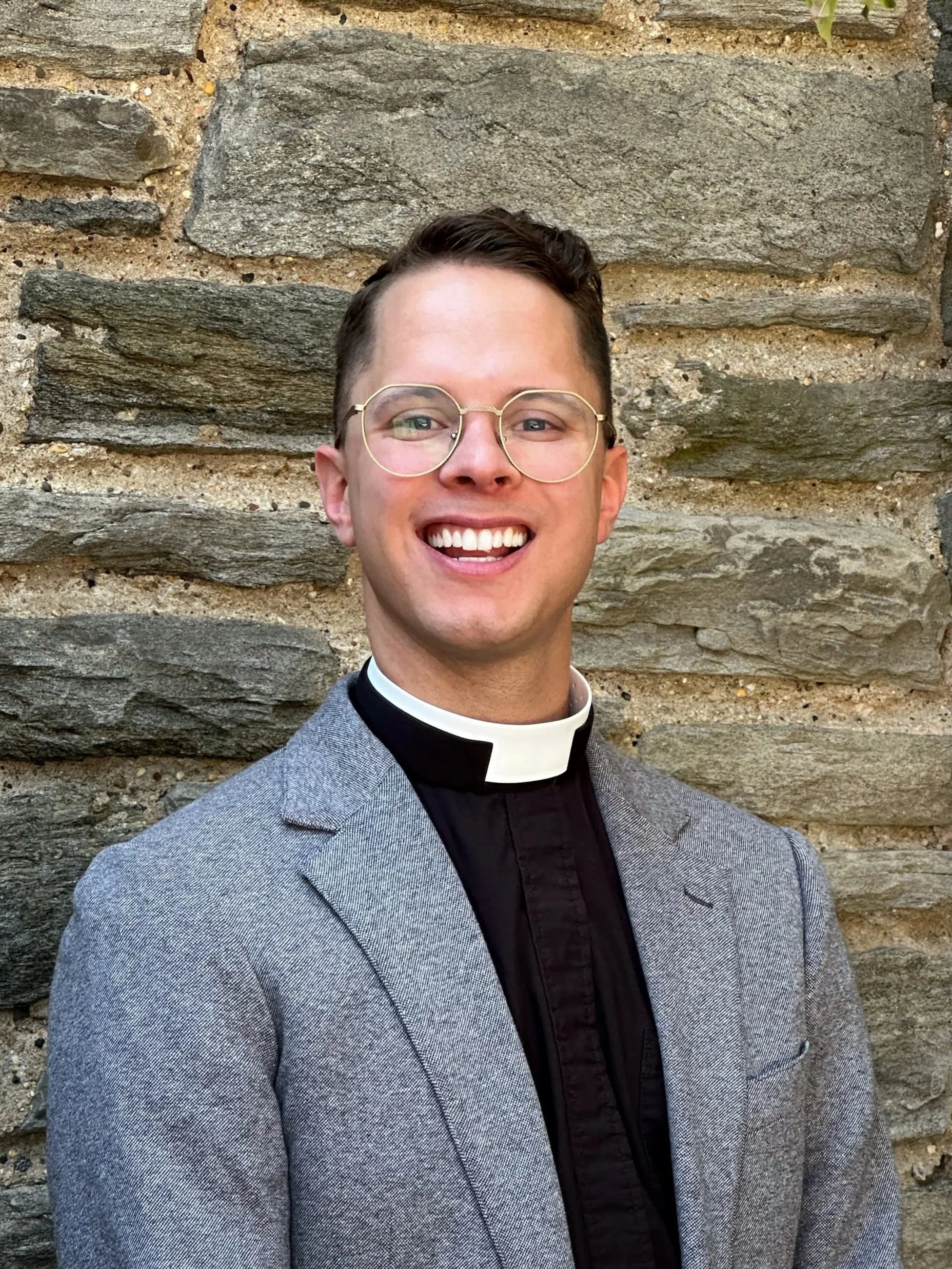 A portrait of a smiling man wearing glasses, a clerical collar, a gray blazer, and a black shirt, standing in front of a stone wall. Priest of the church.