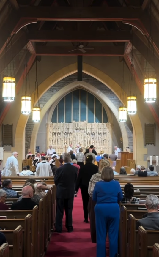 People seated and standing inside a church with wooden pews, chandeliers, and a decorative altar in the background during a service or event.