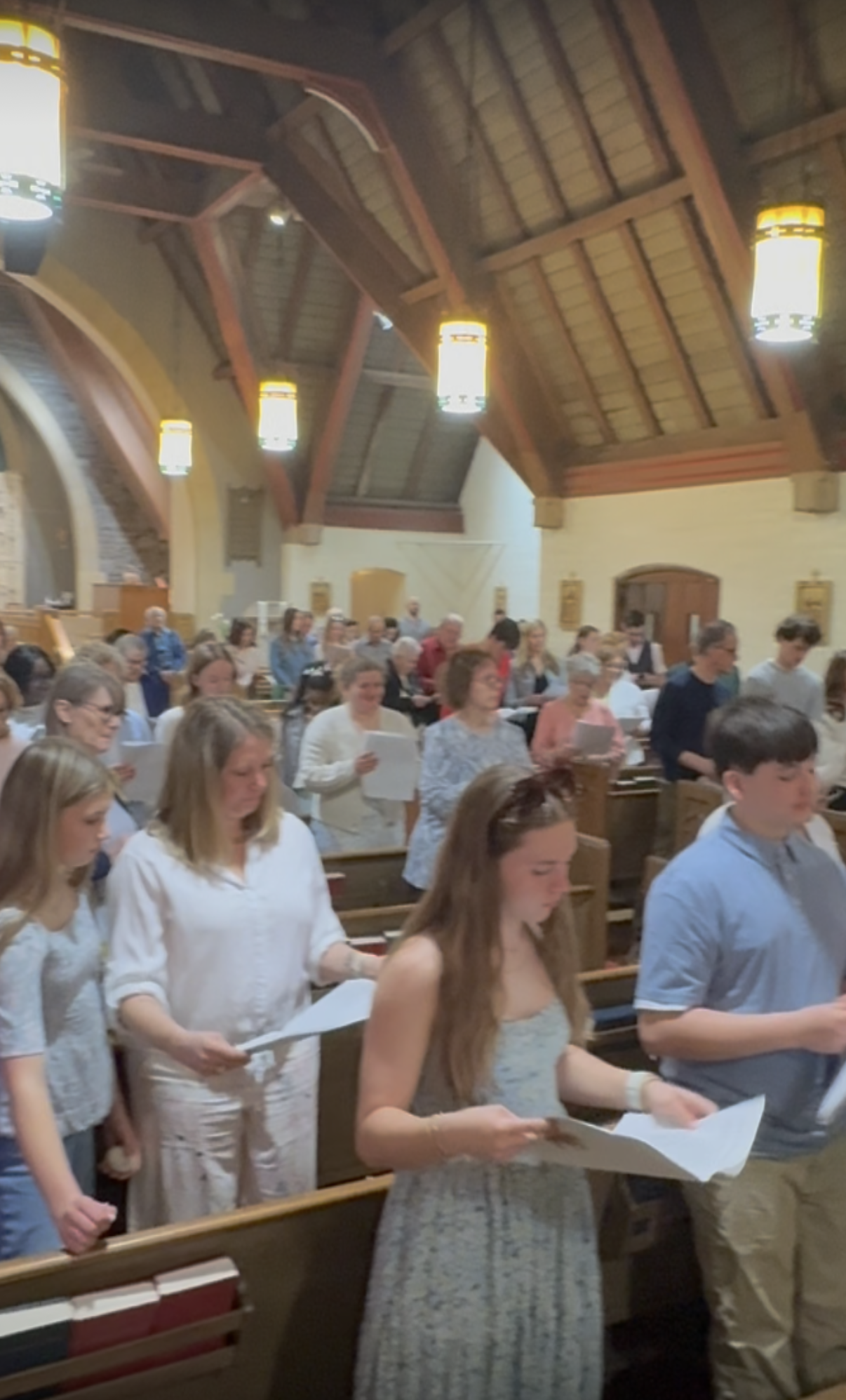 People inside a church standing in pews, holding papers, possibly singing or reading during a service. The church has wooden beams, a high ceiling, and hanging lights.