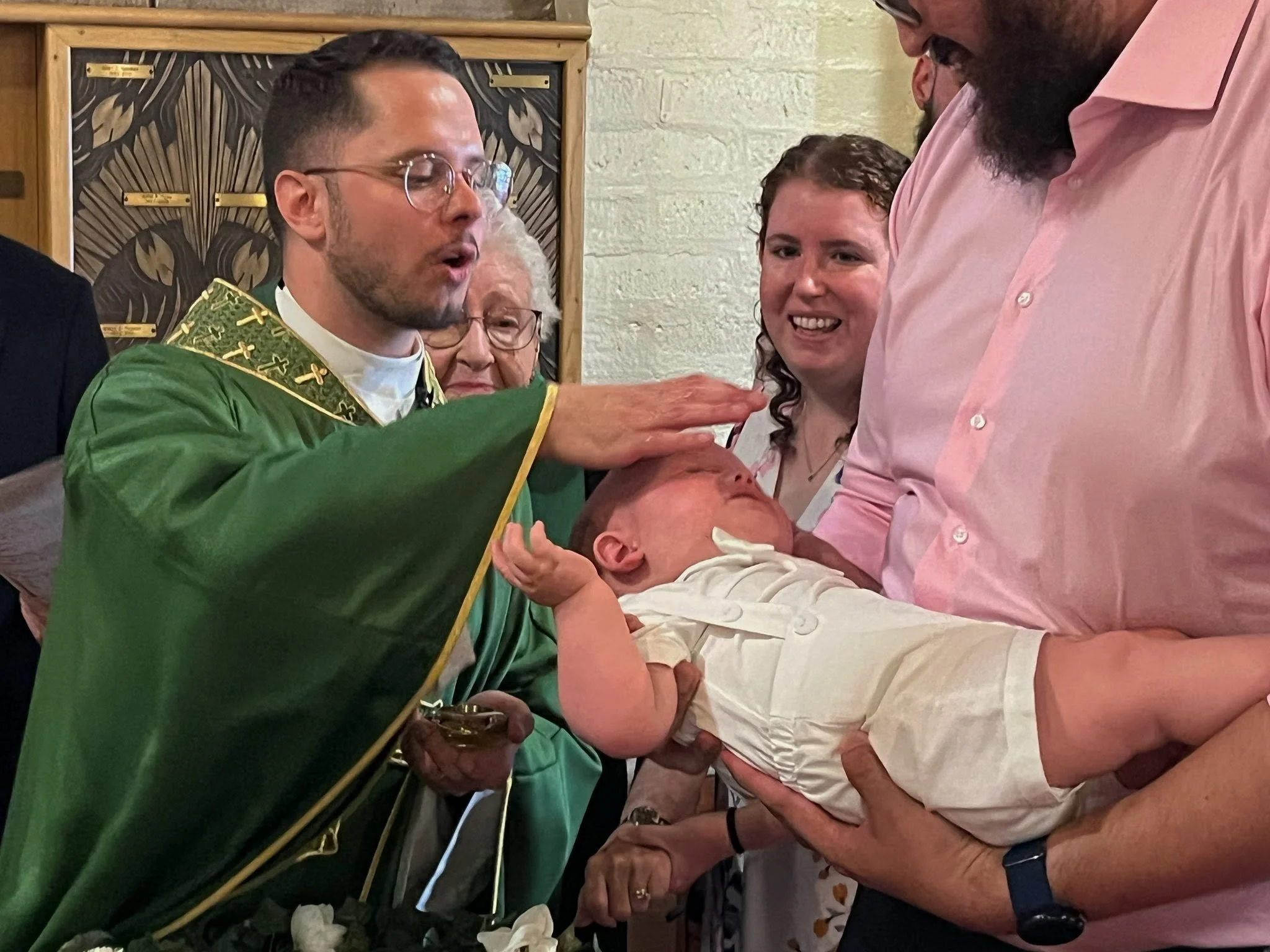 A group of people are gathered around a baby during a baptism. The Priest is dressed in green vestments. The other adults are smiling and watching attentively.