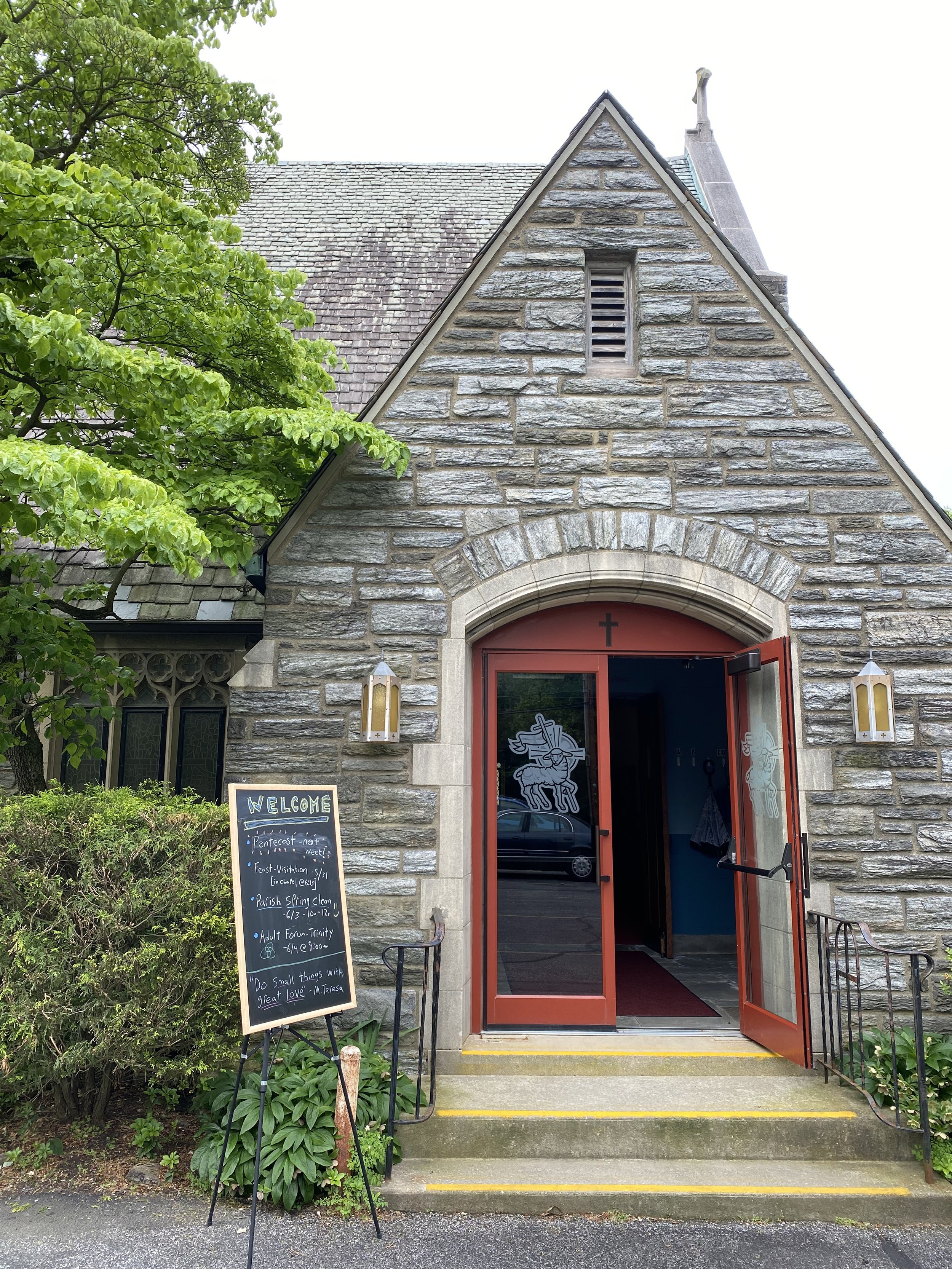 The image shows the entrance of a church with a stone facade and a red door. There are steps leading to the door, flanked by two lantern-style lights. A blackboard with a welcome message and event notes stands on the left side of the entrance, surrounded by greenery and bushes. The church's roof is visible, with a small vent or window. Part of a tree with green leaves is on the left side of the image.
