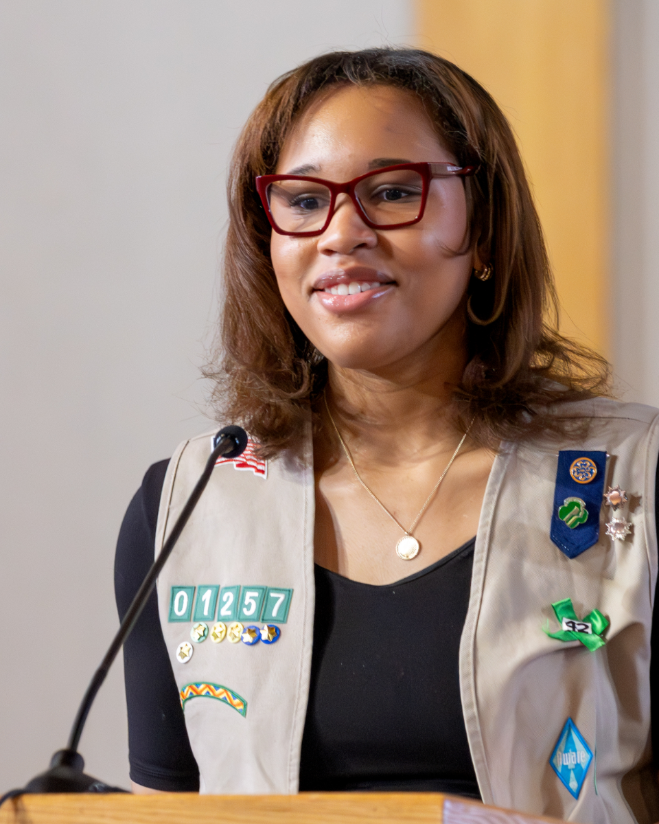 A woman with shoulder-length brown hair, red glasses, and a tan Girl Scout vest with badges and patches, speaking at a podium.