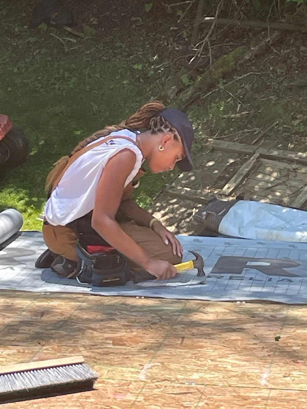 A person with long braided hair wearing a cap, white sleeveless shirt, and brown pants is kneeling on a patterned mat outdoors. They are handling a yellow object, with a backpack nearby and a broom in the foreground. The background shows green foliage and a rustic wooden fence.