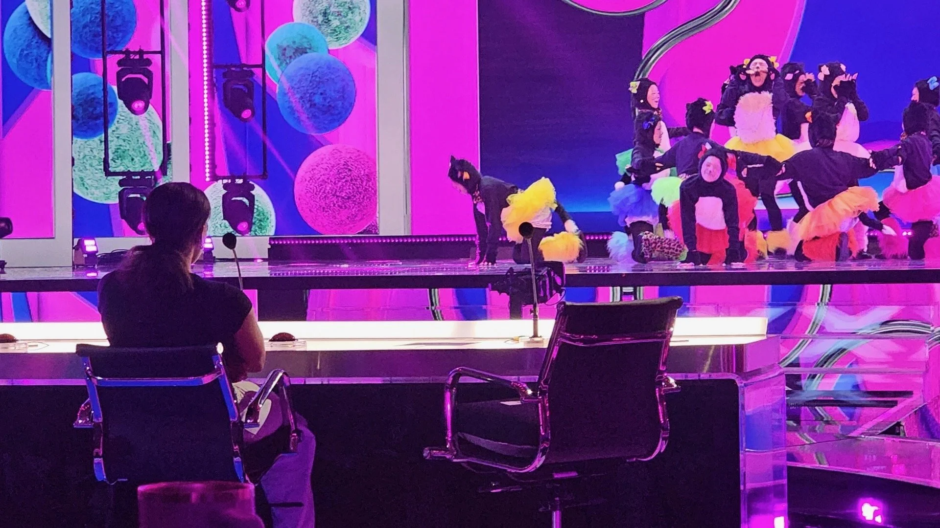A young girl in a bear costume performs on stage with other children dressed as animals, while a woman watches from behind the glass in a dark room with pink and purple lighting.