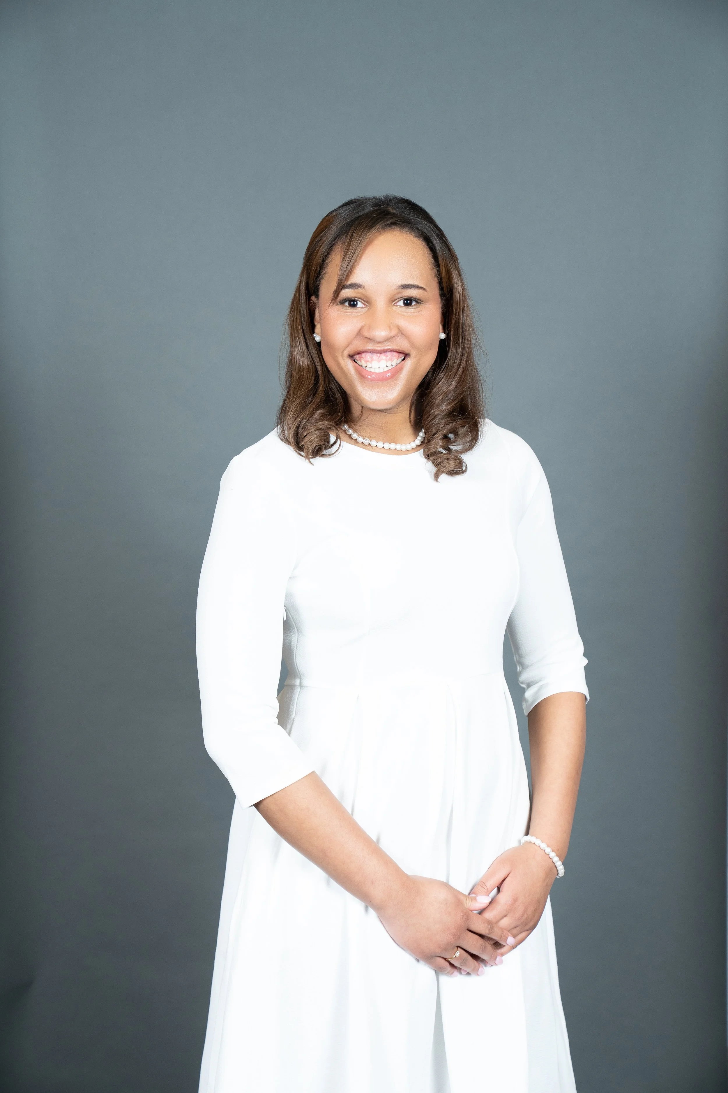 A smiling woman wearing a white dress, pearl necklace, pearl earrings, and a bracelet, standing against a neutral gray background.