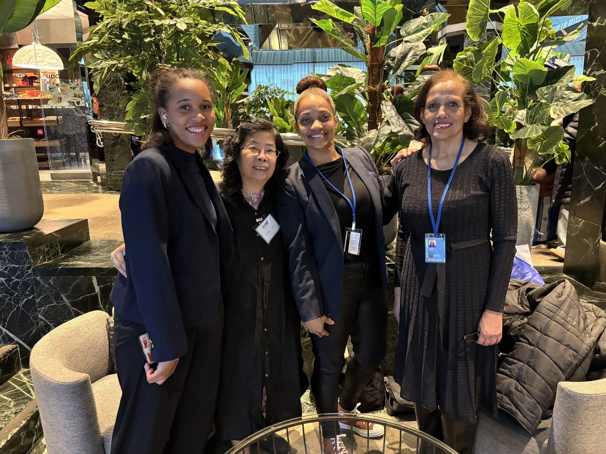 Four women standing together in a lobby, smiling at the camera. They are surrounded by large green plants and dark marble decor. They are wearing professional attire, and some have conference badges on lanyards around their necks.