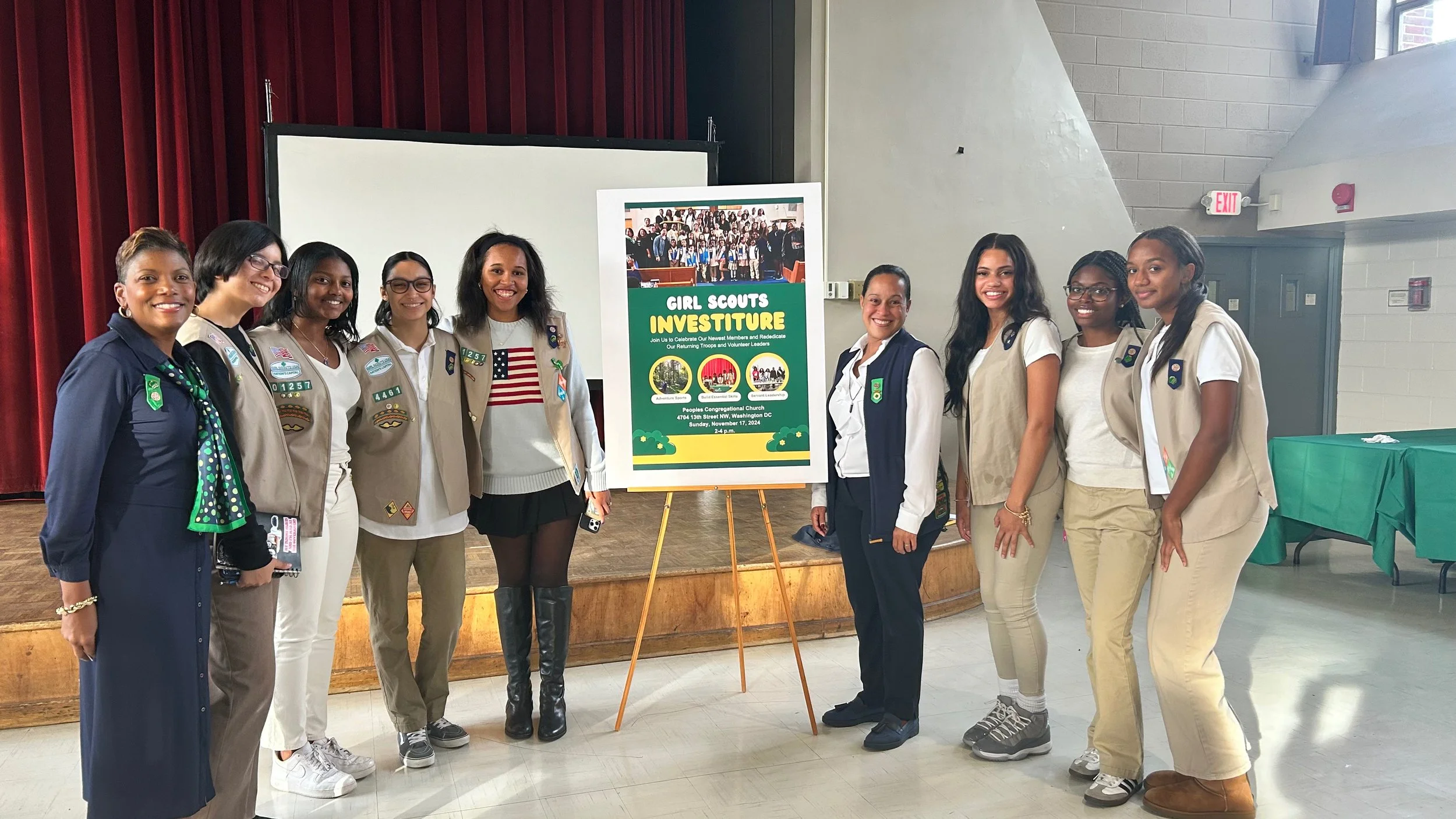 Group of girl scouts indoors, standing on a stage in front of a sign for a girl scouts investiture ceremony, with a red curtain and a white wall in the background.