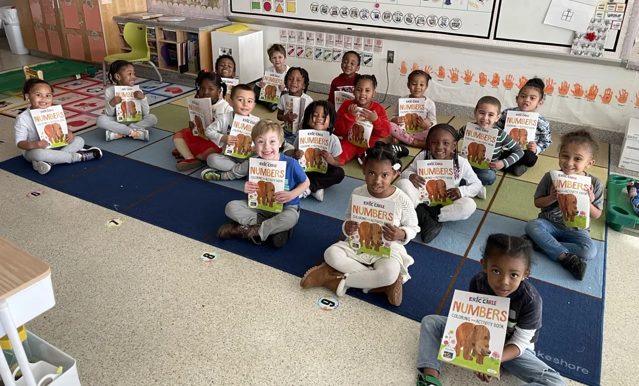 Group of young children sitting on a classroom carpet, each holding a coloring book titled 'Eric Carle Numbers' featuring a bear on the cover.