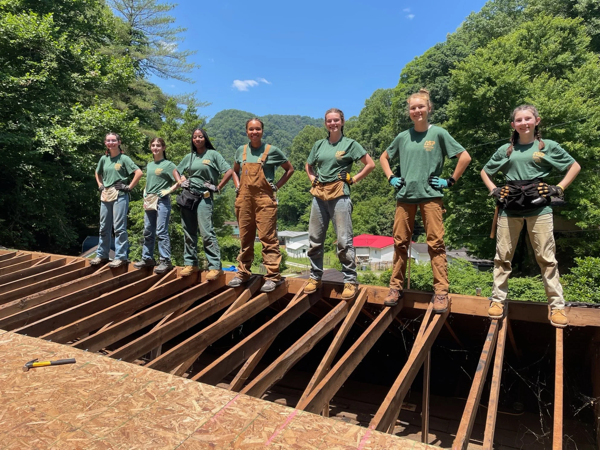 Seven young women standing on a wooden deck frame outdoors, wearing green T-shirts and work gloves, with a lush green hillside and blue sky in the background.